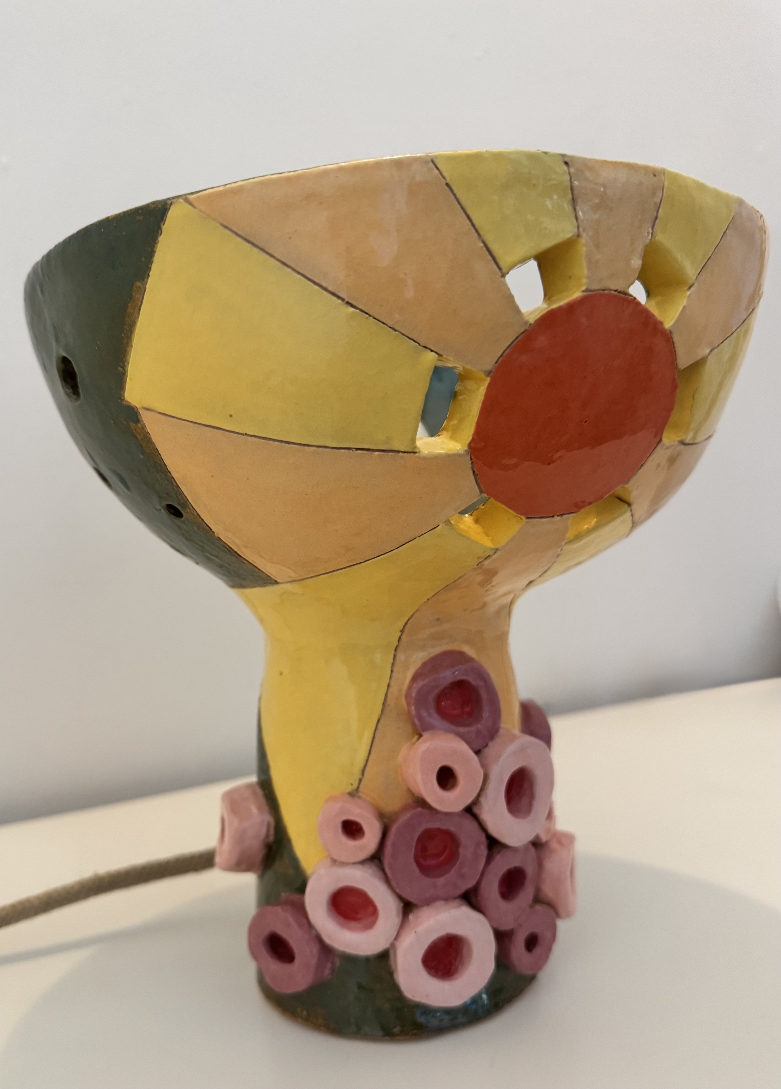Colorful ceramic sculpture of a wine glass with a cluster of pink grapes at the base. The bowl of the glass is decorated with orange, yellow, and black sections, resembling a geometric pattern.