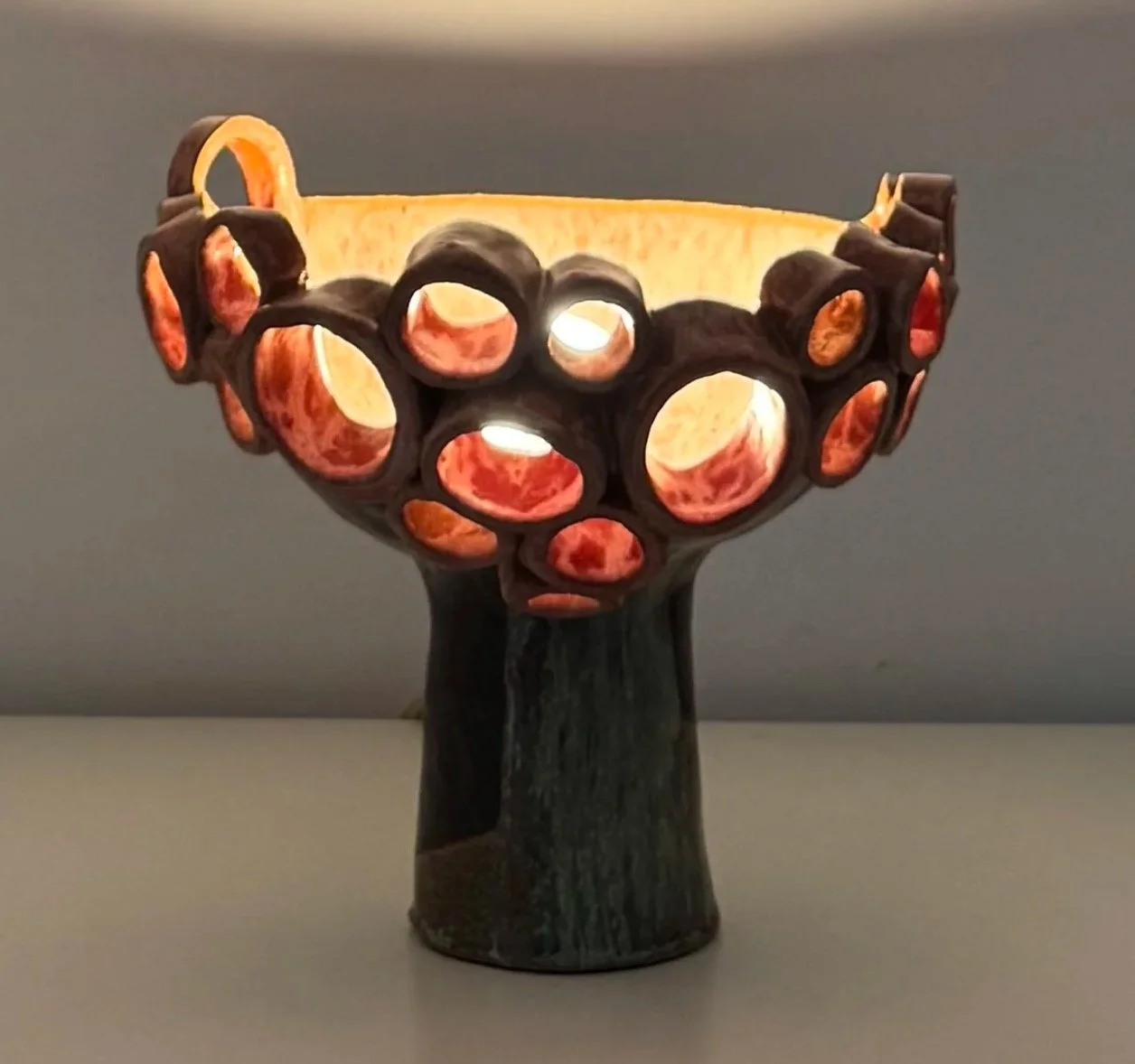 Decorative ceramic bowl with a branch-like base and circular cutouts, illuminated from inside, against a gray background.