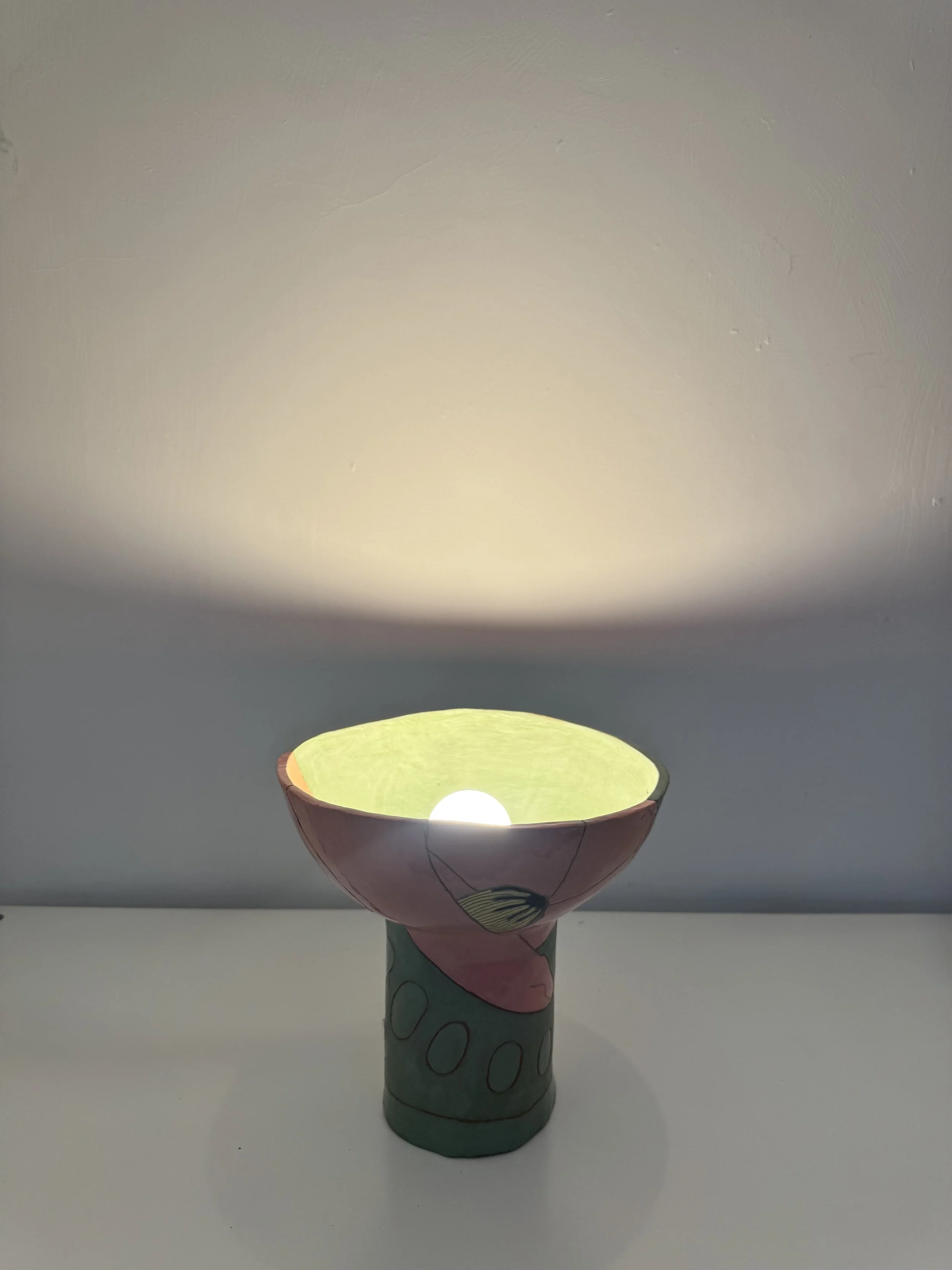 A decorative ceramic or clay lamp with a hand-painted design of what appears to be a bird, placed on a white surface against a plain light-colored wall. The lamp is lit from within, casting a warm glow.