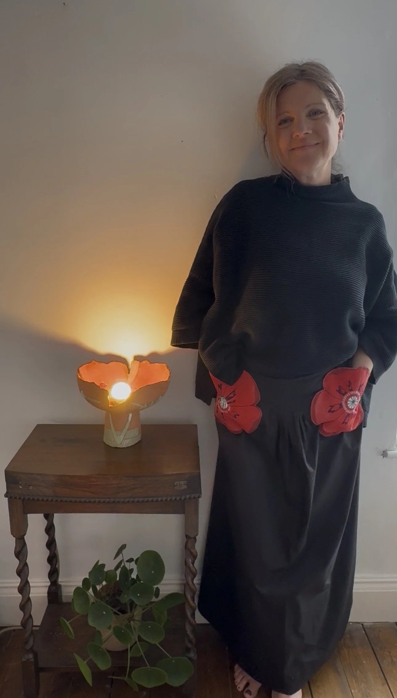 A woman with short hair standing beside a wooden table with a lit candle in a decorative holder. She is wearing a black top with red flower designs at the waist, and wide black pants. A green potted plant with round leaves is on the floor in front of the table, and a soft light glows in the background.