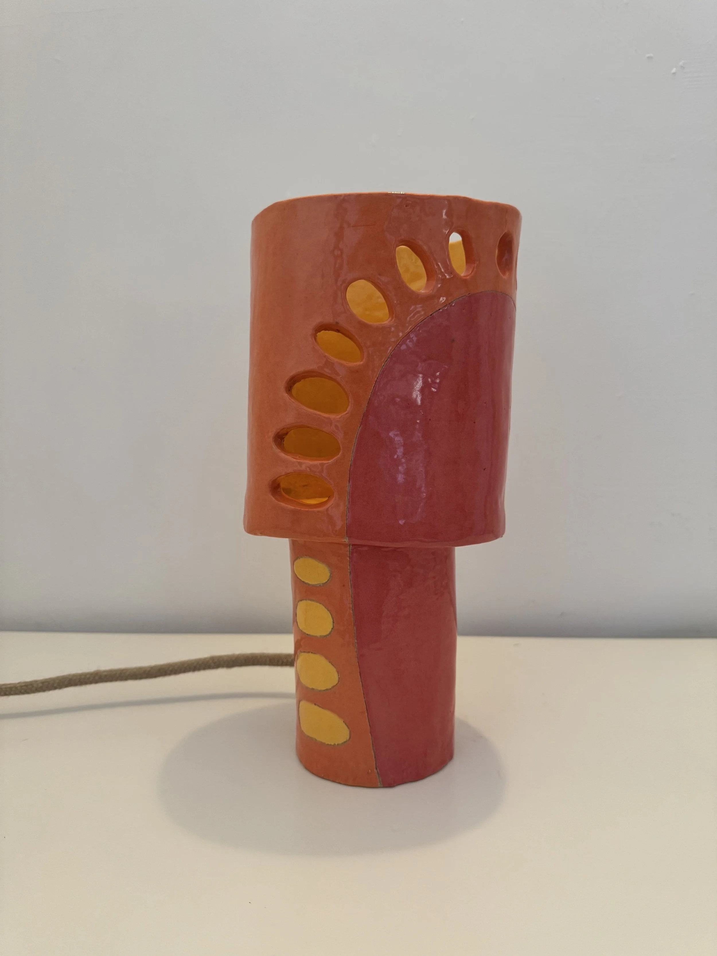 Colorful lamp with oval cutouts on an orange and pink gradient surface, standing on a white surface against a plain white background.