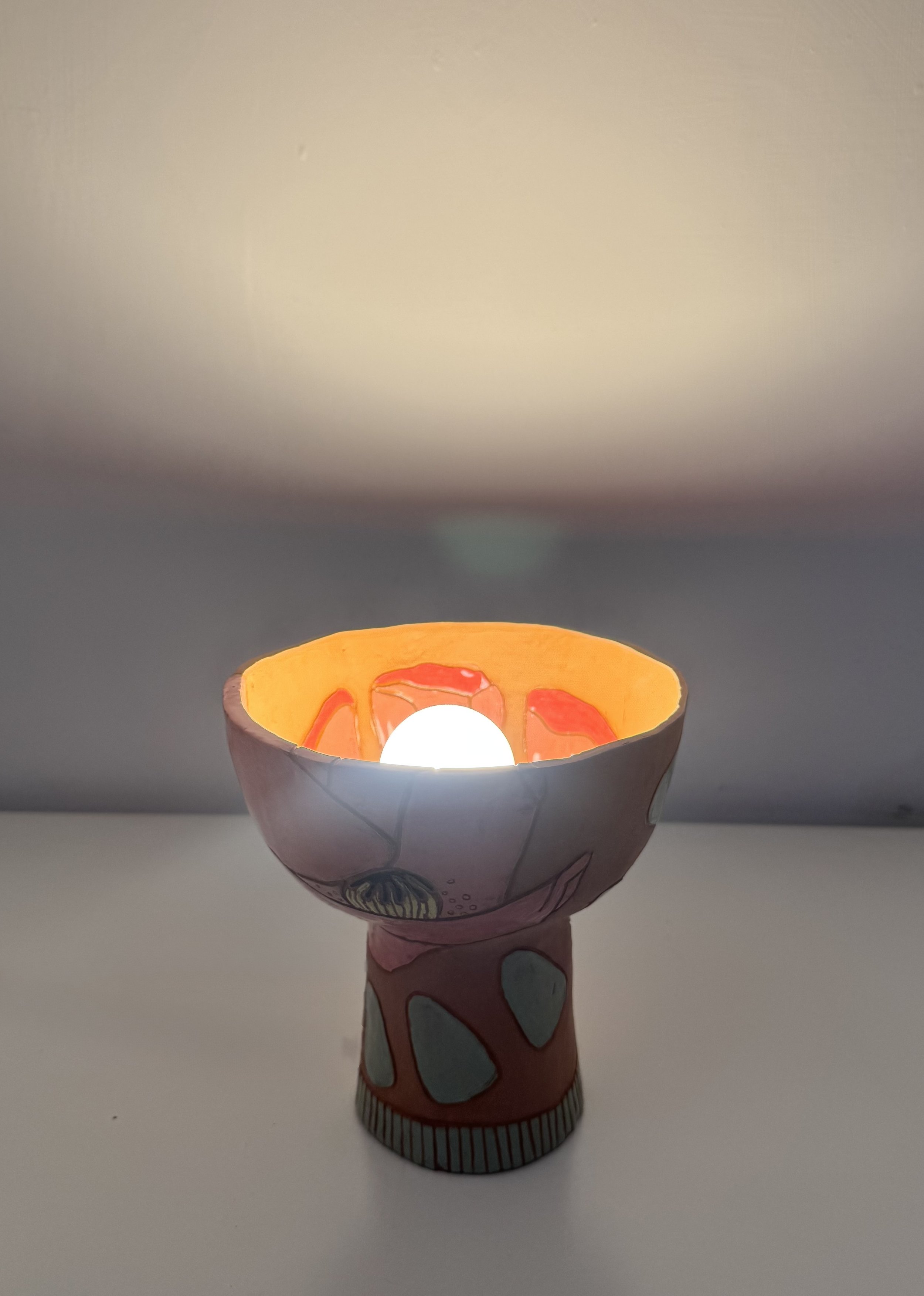 A lit paper lamp in the shape of a mushroom with a decorated cap and stem, placed on a white surface against a plain background.