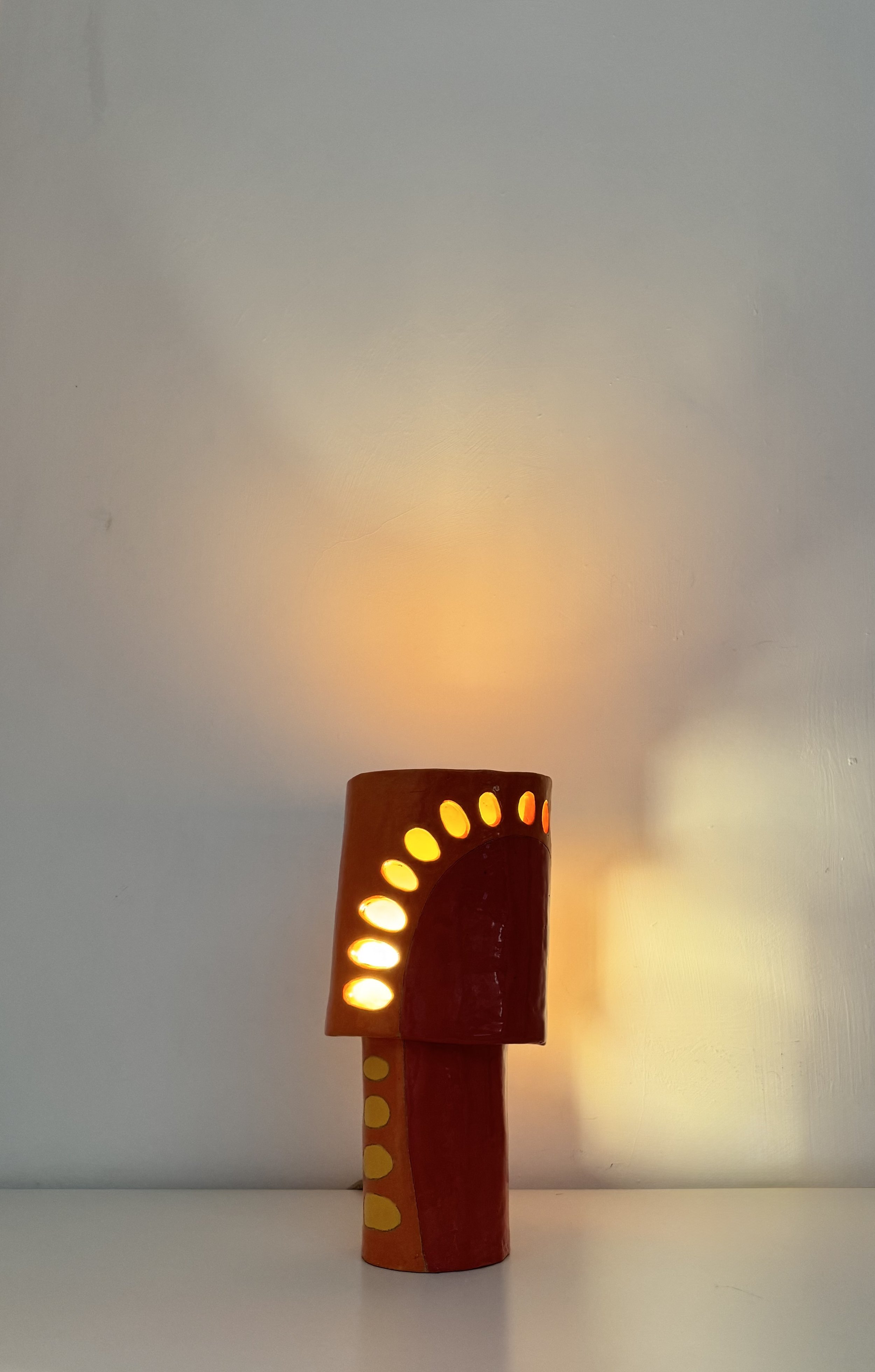 Decorative cylindrical lamp with yellow oval cutouts, emitting warm light, placed on a white surface against a plain white wall.