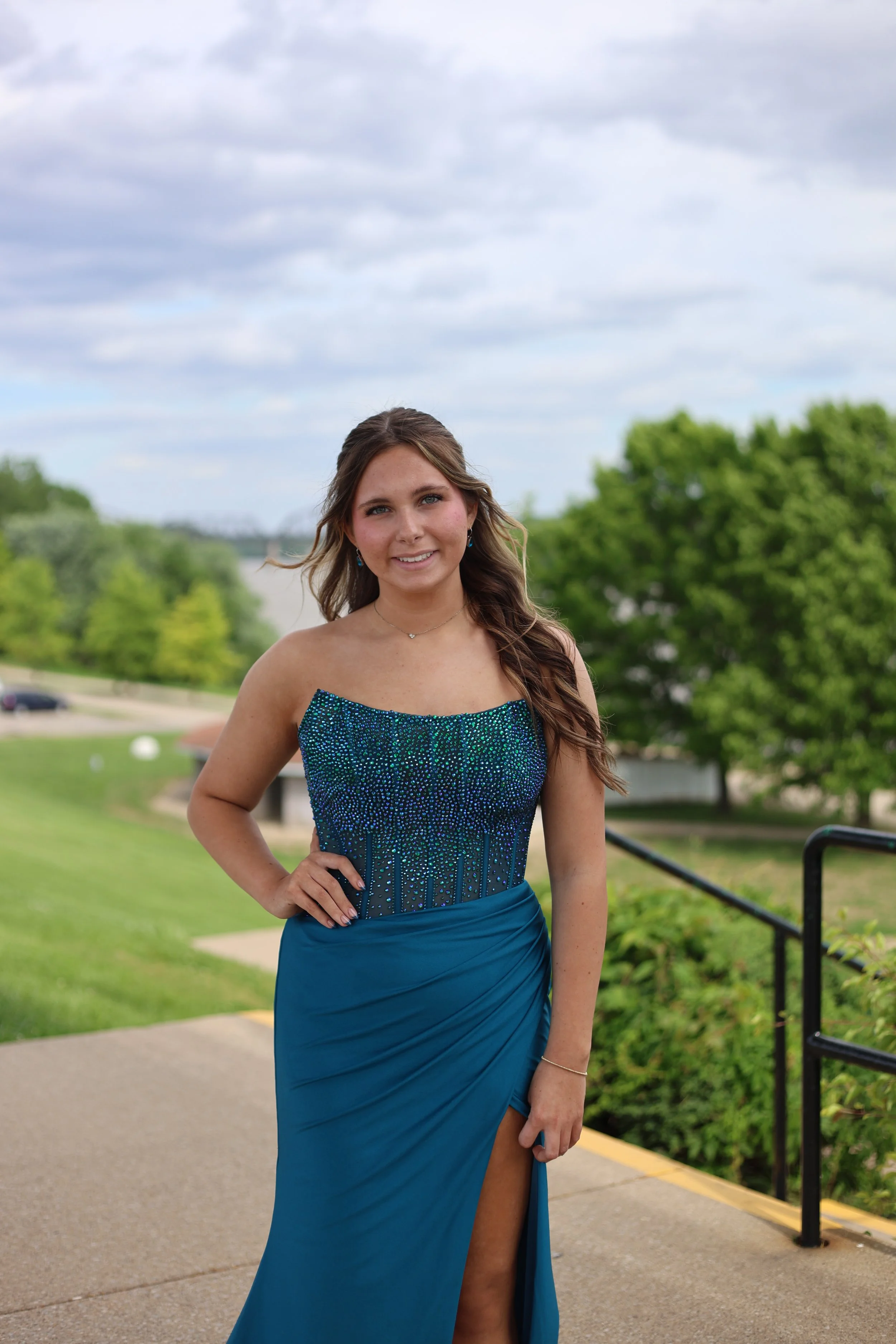 Young woman wearing a blue, embellished strapless dress with a high slit, standing outdoors on a cloudy day with greenery and water in the background.
