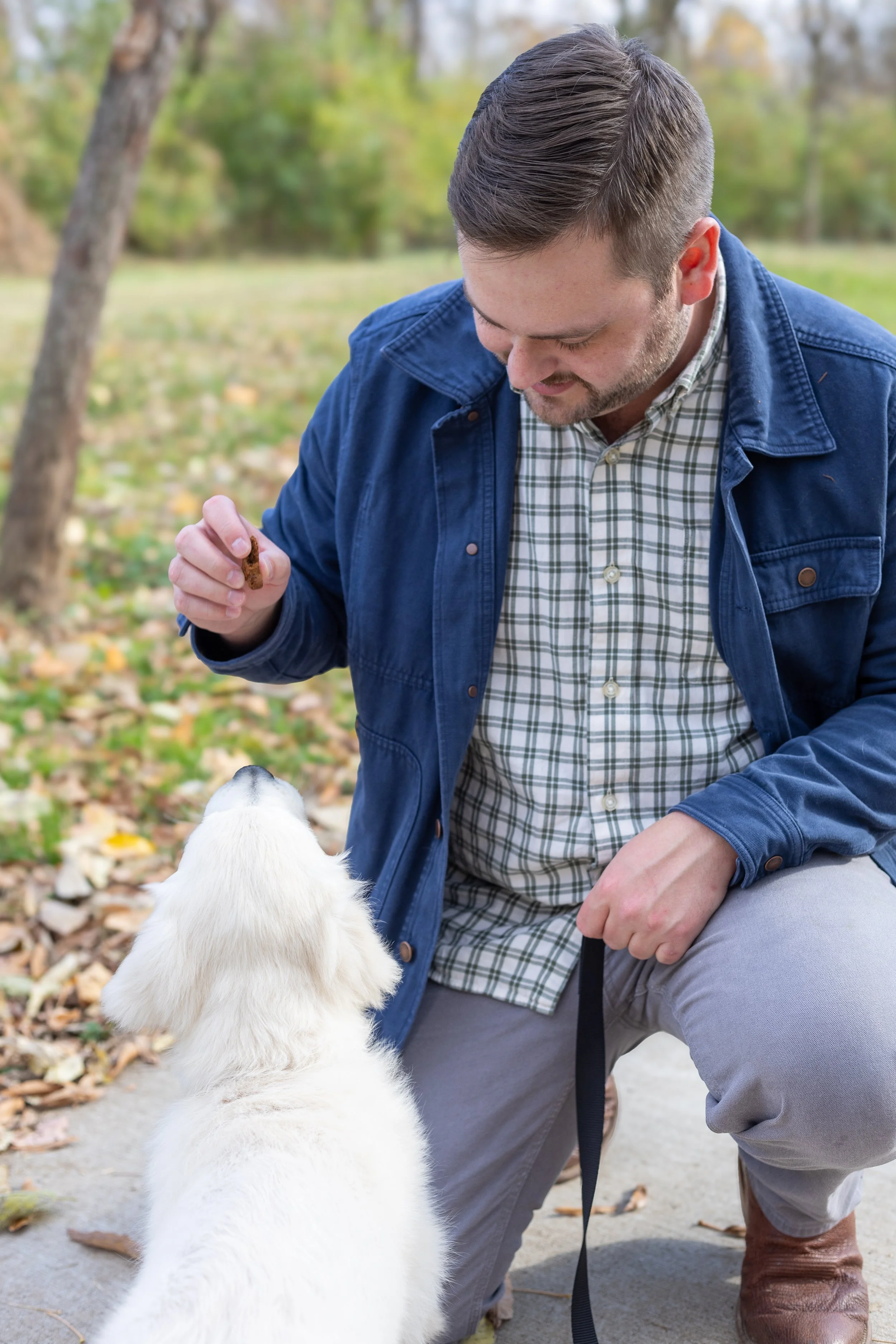 A man with a plaid shirt and blue jacket kneels on the ground, holding a treat, and interacts with a white puppy in an outdoor park with autumn leaves.
