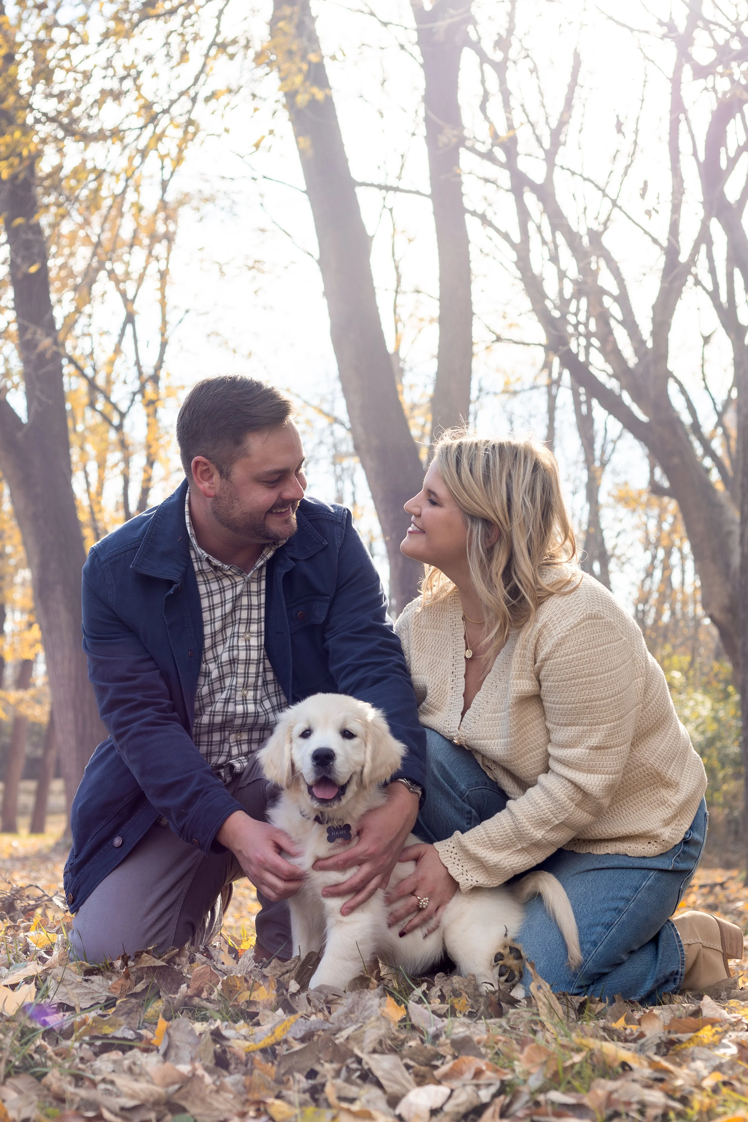 A couple and a woman smiling and playing with a golden retriever puppy outside in a park with fallen autumn leaves and tall trees in the background.
