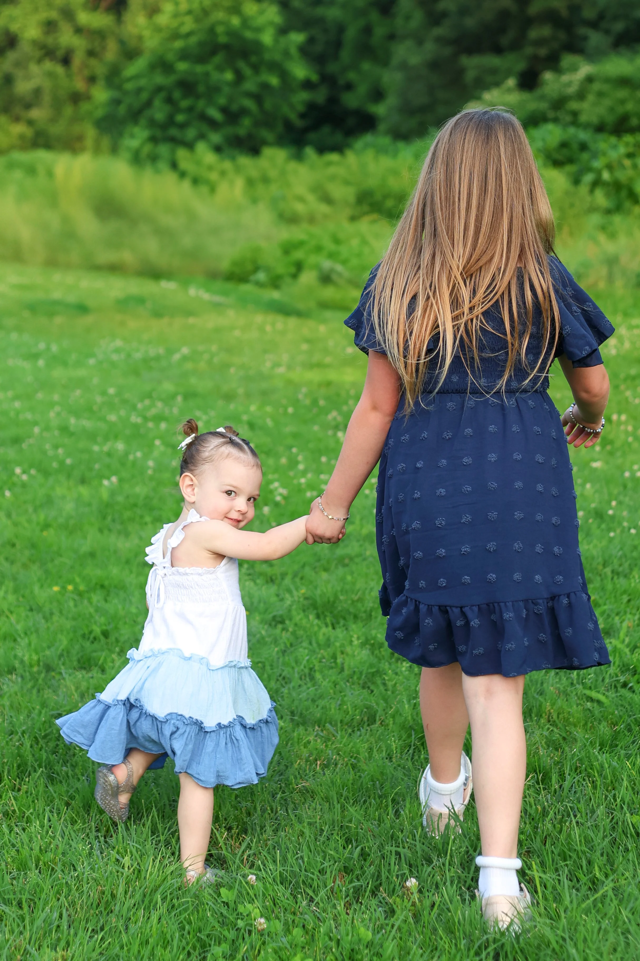 A woman holding hands with a young girl and walking through a grassy field as trees and greenery are visible in the background.