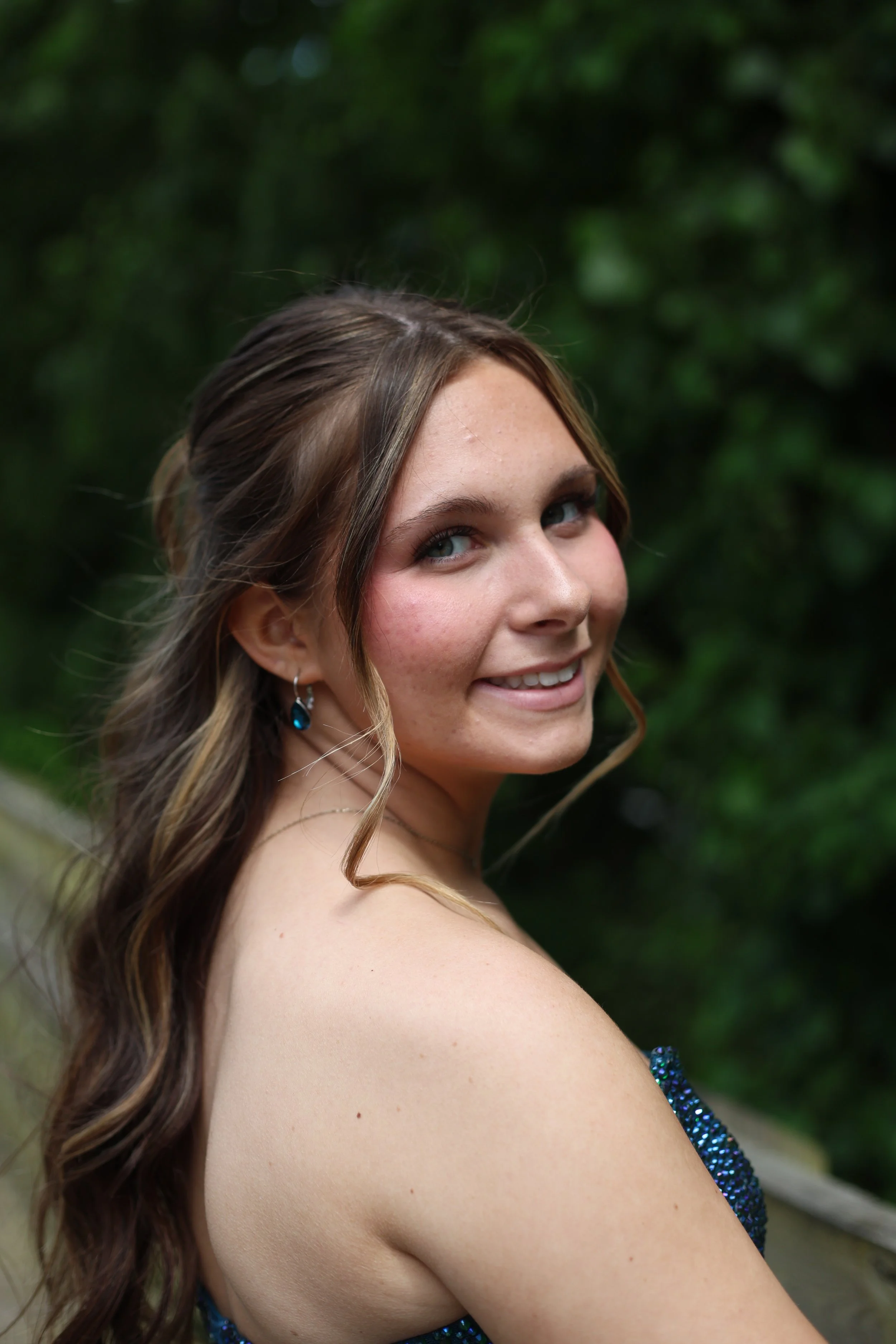 A young woman with long, wavy brown hair and blue eyes smiling outdoors. She is wearing a strapless, sparkly blue dress and blue earrings, with green foliage in the background.