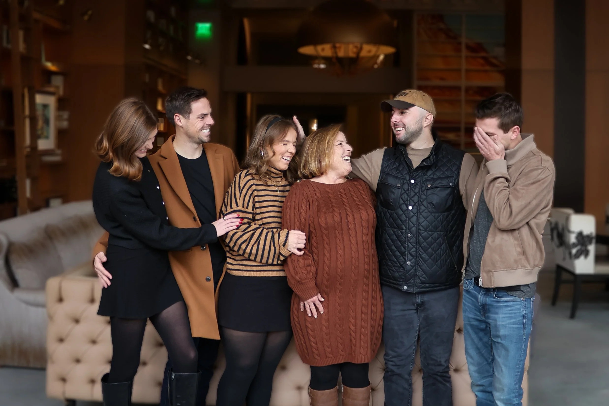 Six people are gathered together, smiling, and enjoying each other's company in a warmly lit room. They are standing in front of a beige tufted sofa, with a bookcase and a large lamp in the background. The group appears to be engaged in a joyful conv