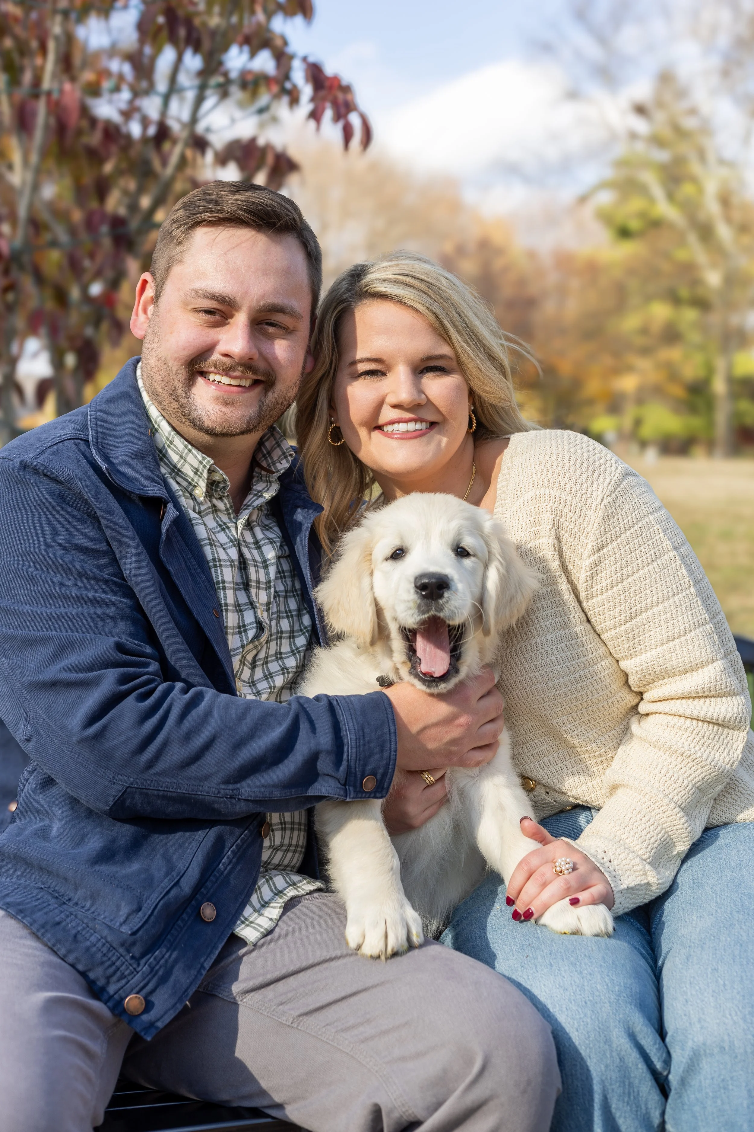 A smiling man and woman sitting outdoors, holding a Golden Retriever puppy, with autumn trees in the background.