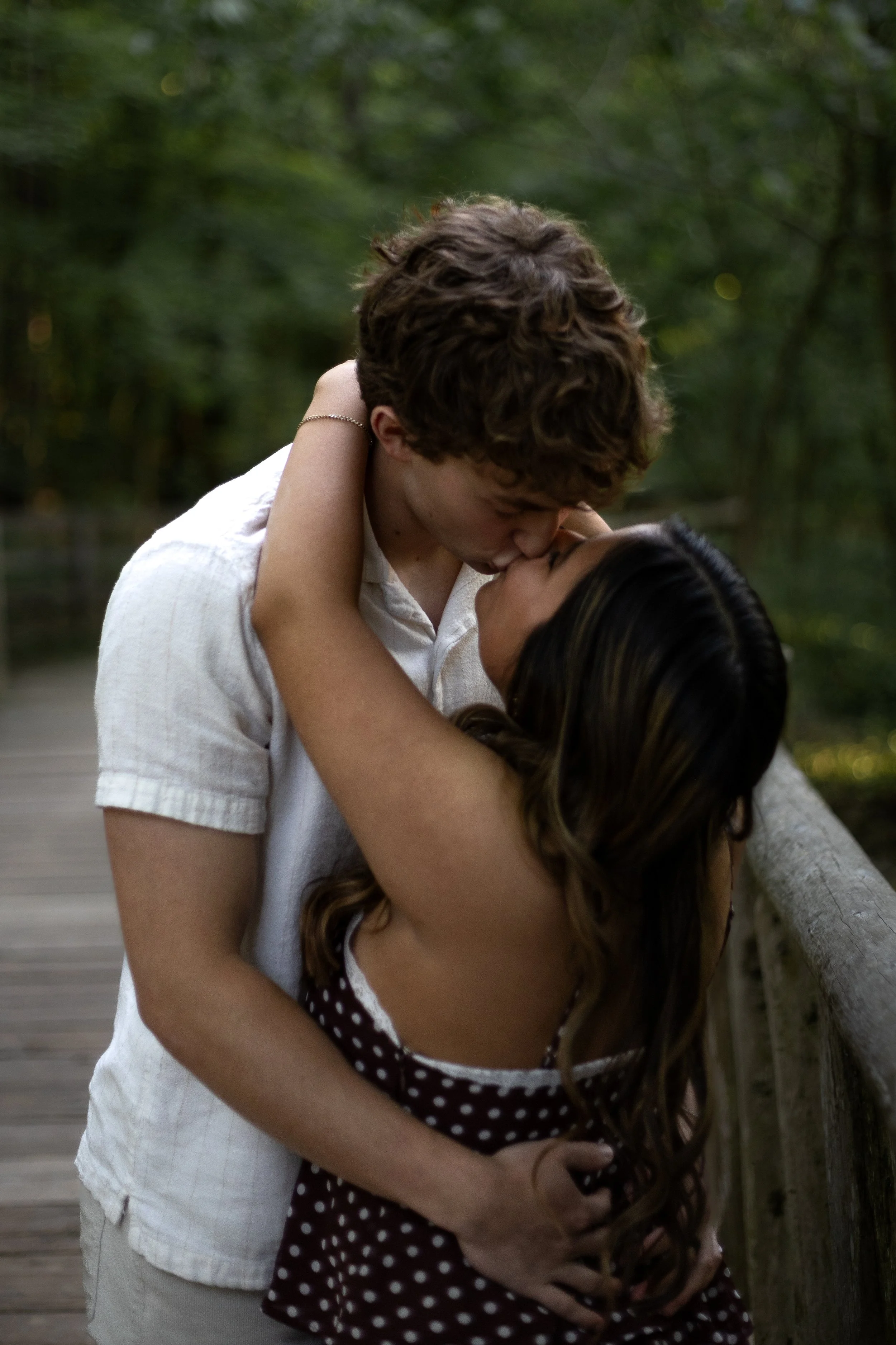 A young couple sharing a kiss outdoors on a wooden bridge with greenery in the background.