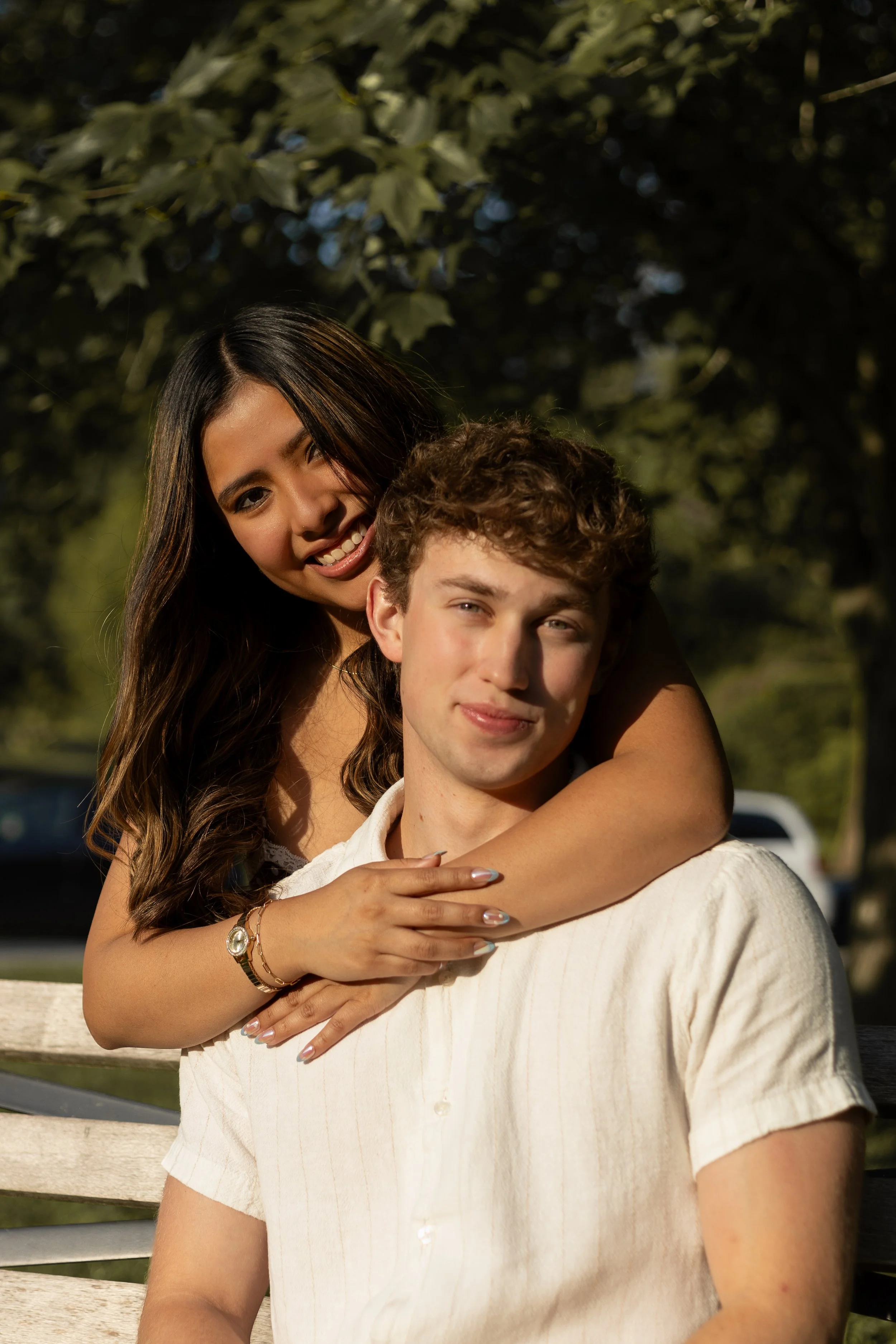 A young woman with long dark hair smiling with her arm around a young man with curly hair, outdoors with green trees in the background.