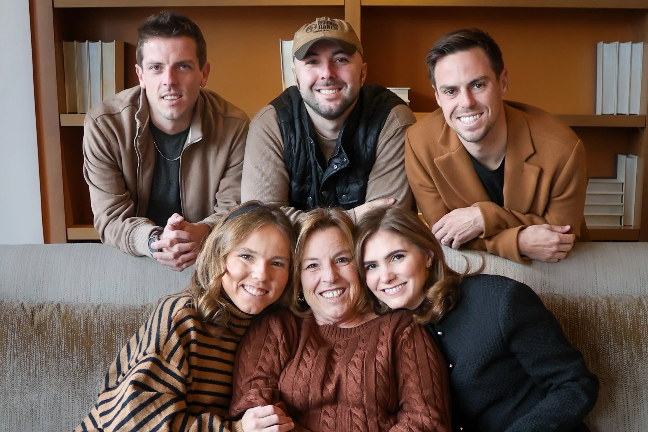 A group of six smiling people, three men and three women, posing together indoors in front of wooden bookshelves.