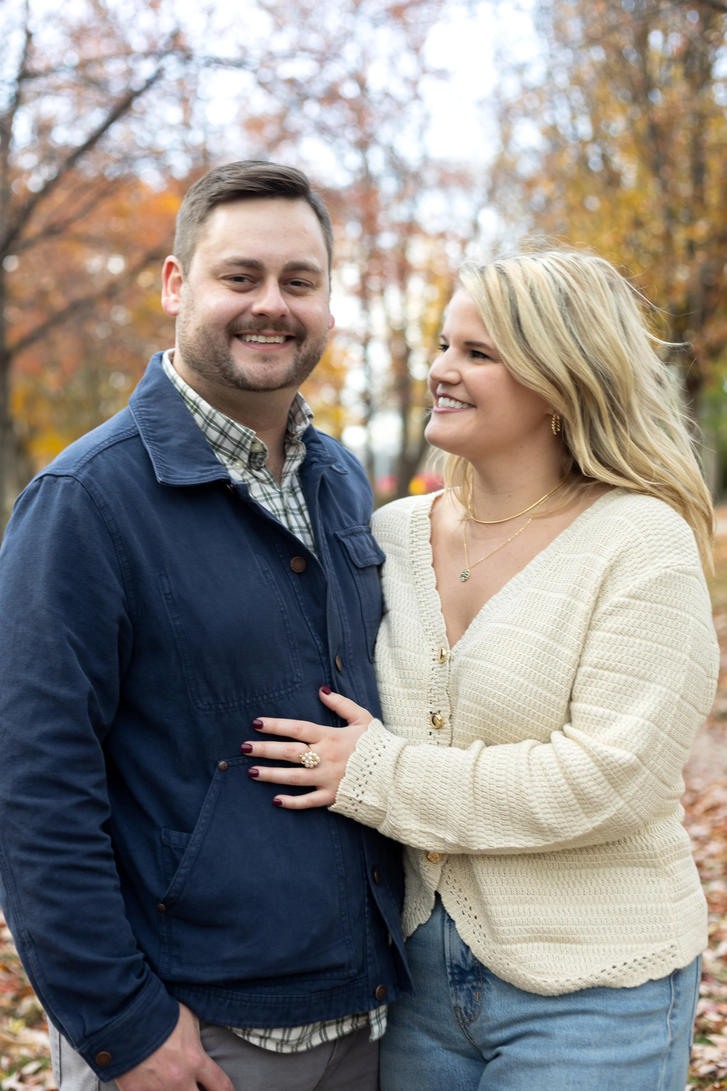 A smiling man and woman standing close together outdoors during autumn with colorful fall foliage in the background.