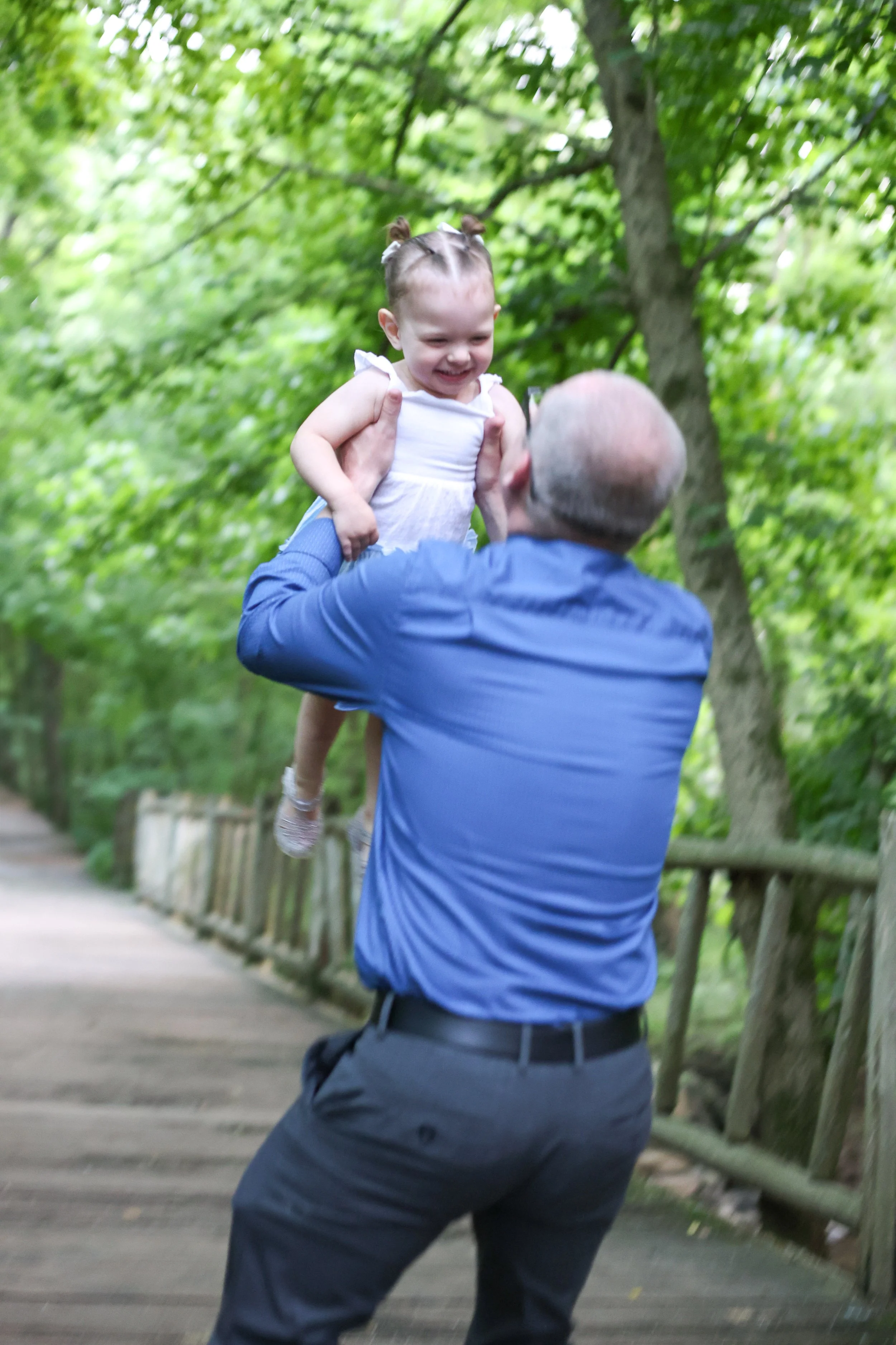 A man in a blue shirt holding a young girl in a white dress up in the air on a wooden path surrounded by trees.