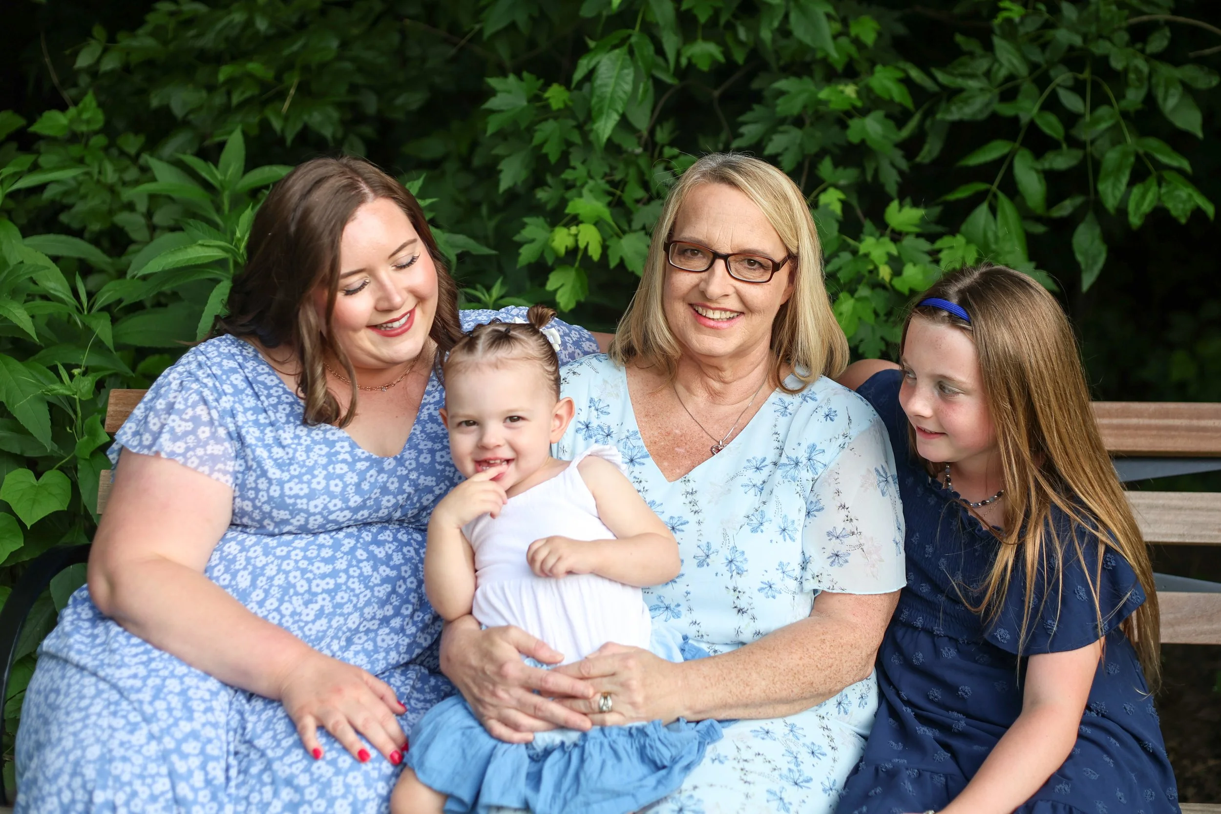 Four generations of women and two young girls sitting on a wooden bench outdoors, surrounded by green foliage, smiling and enjoying each other's company.