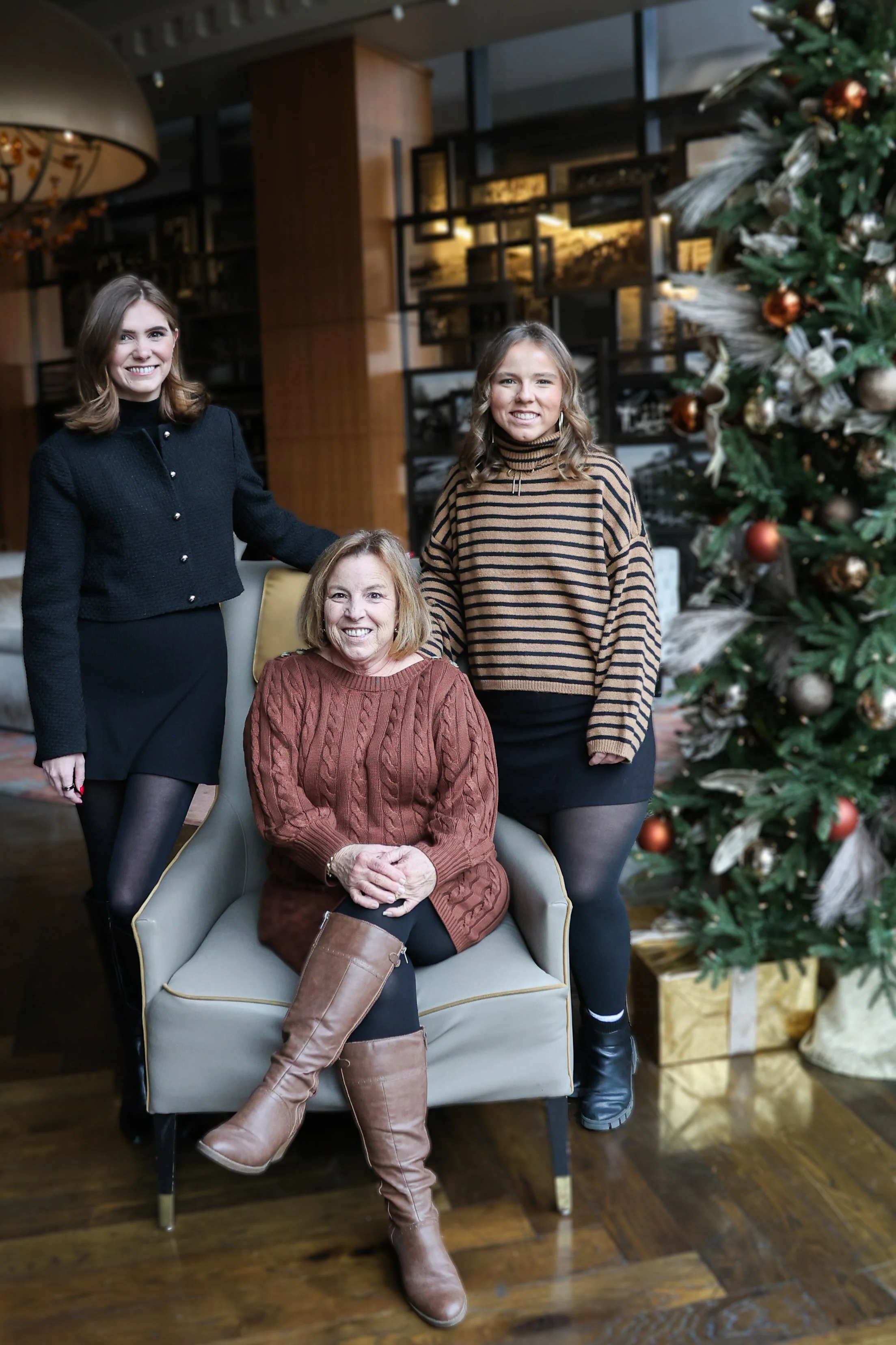 Three women posing by a Christmas tree inside a warmly decorated room. One woman is seated in a chair, wearing a brown cable-knit sweater, brown knee-high boots, and black tights. The other two women stand beside her, smiling, with one in a black bla
