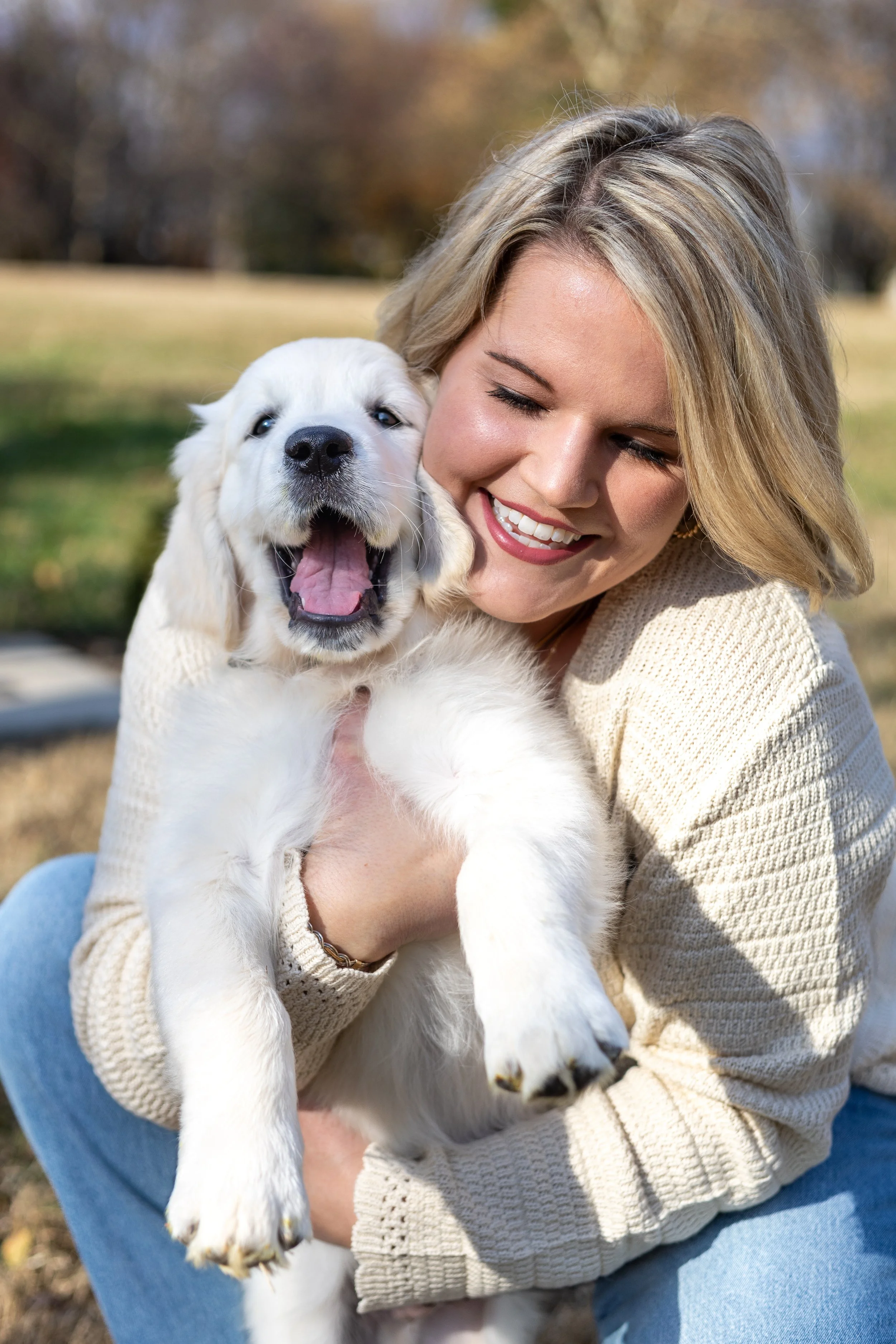 A woman with blonde hair smiling with her eyes closed, holding a happy golden retriever puppy outside in a park with blurred trees in the background.