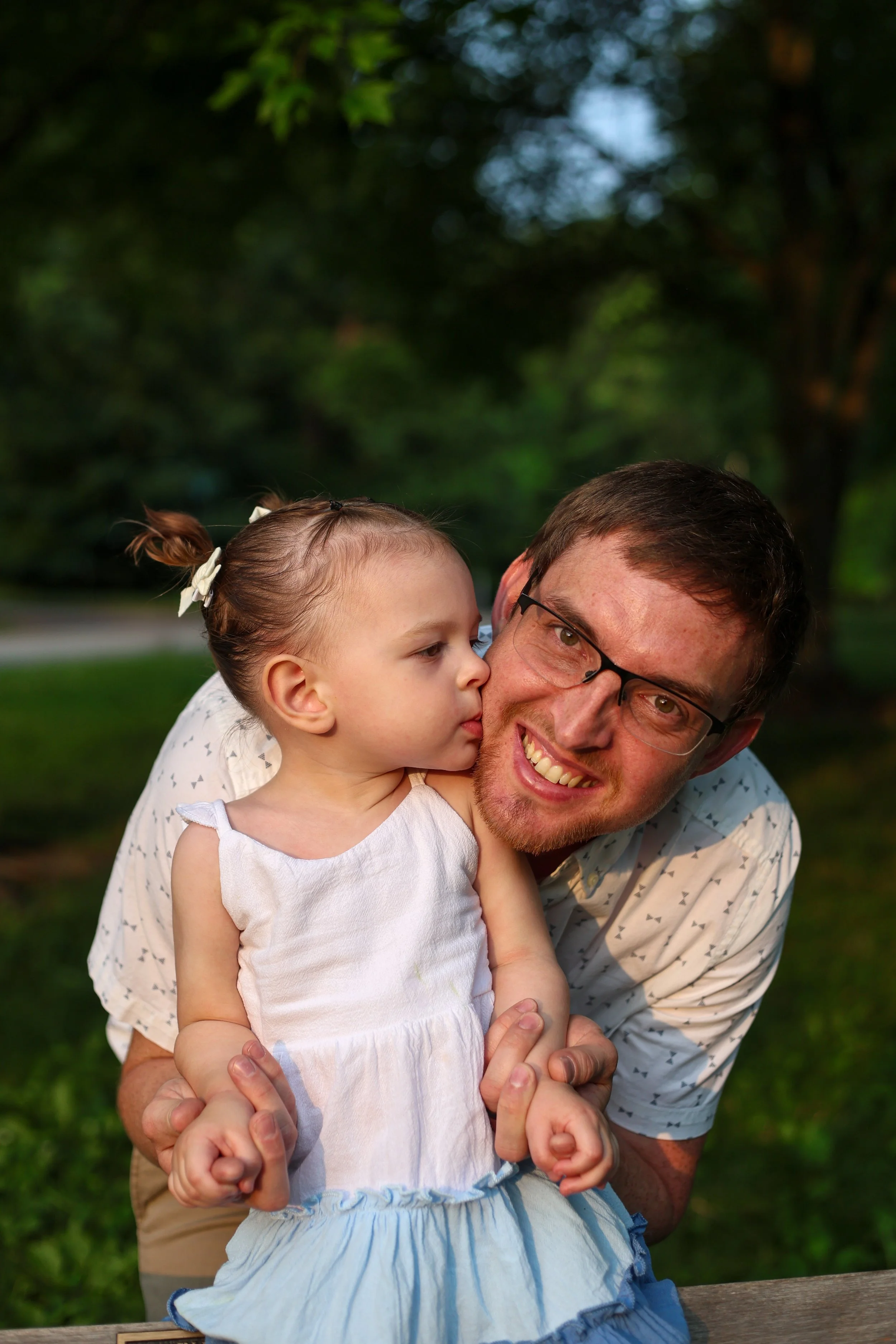 A man with glasses smiling while holding a young girl with a ponytail and a white bow, outdoors with green trees in the background.