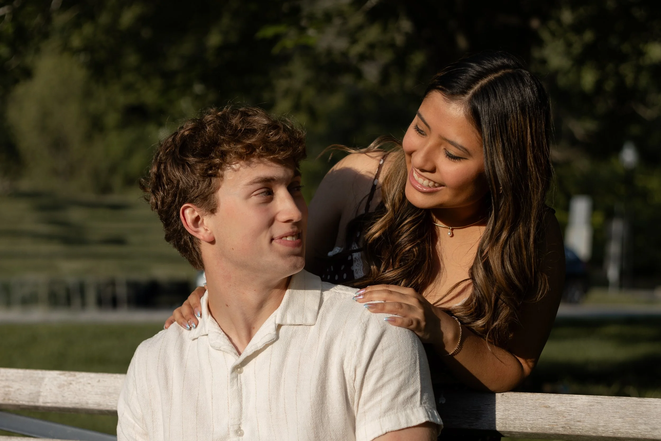 A young man with curly hair sitting on a park bench while a young woman with long, dark hair and a smile leans over to sit next to him, holding his shoulder in a friendly manner. They are outdoors with trees in the background.
