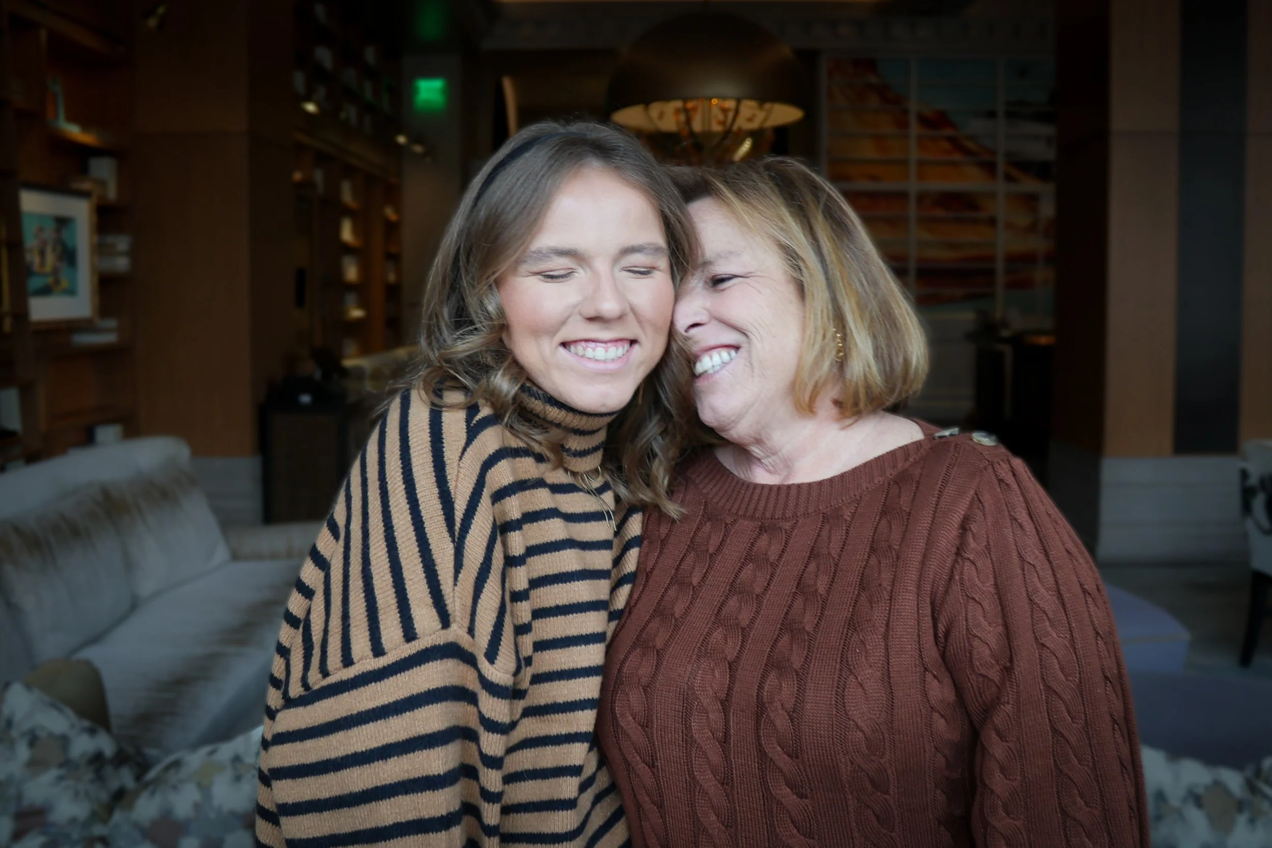 Two women share a warm hug and smile in a cozy, well-lit room with bookshelves and artwork in the background.