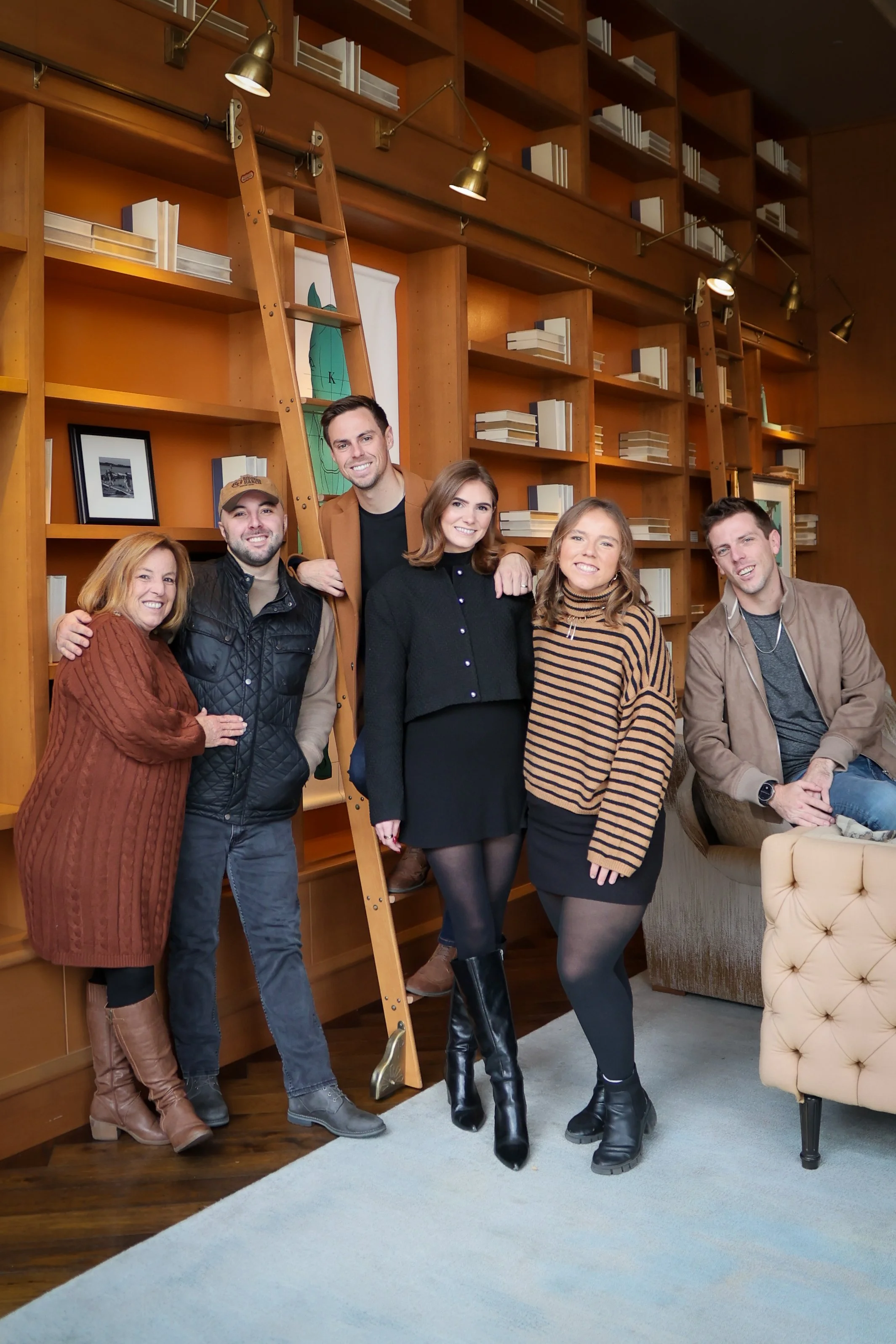 A group of six people posing together in a room with wooden bookshelves and a ladder. They are smiling and dressed in casual attire, some wearing boots, and standing close to each other.