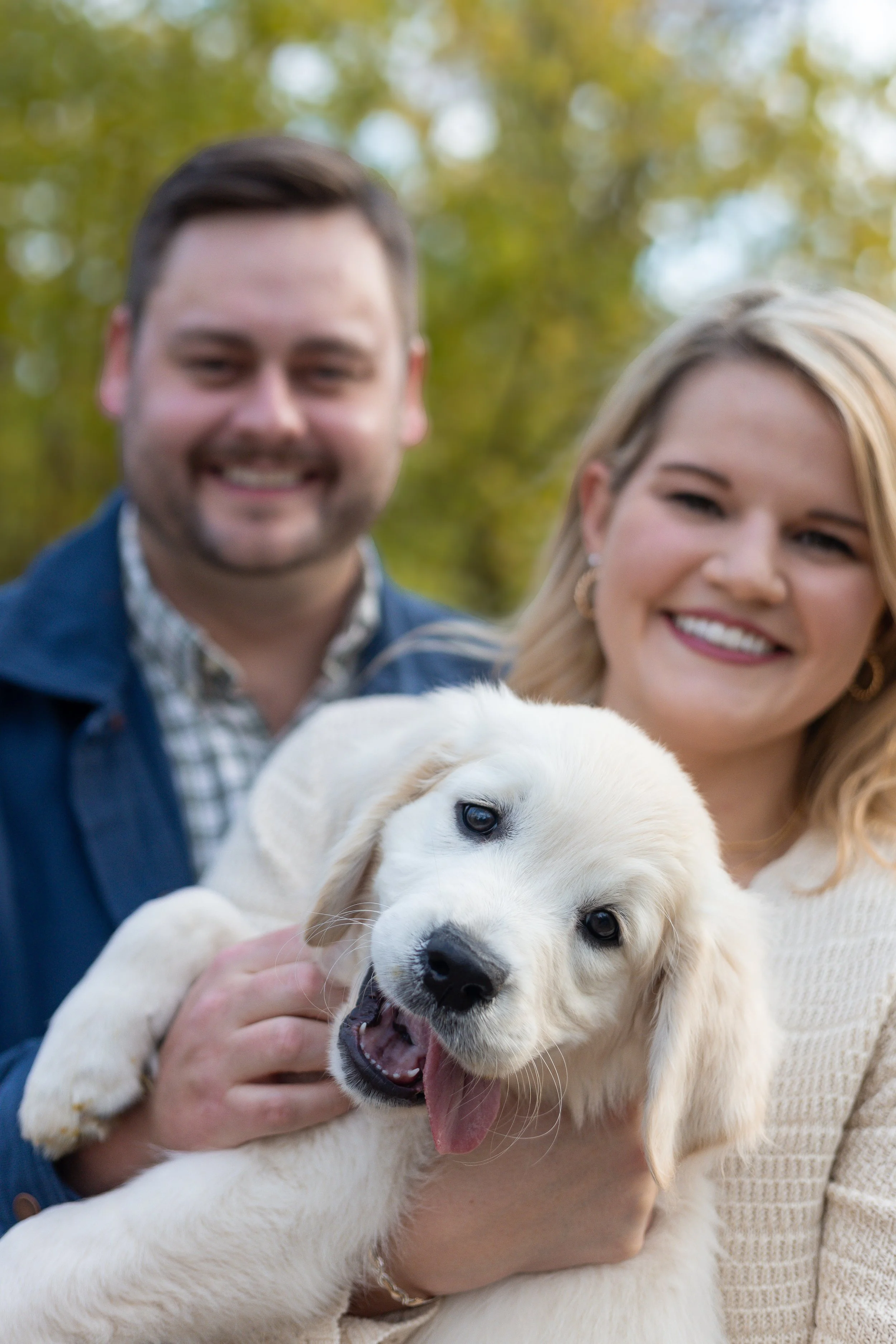 A woman holding a happy Golden Retriever puppy, with a man smiling in the background, outdoors with blurred trees.