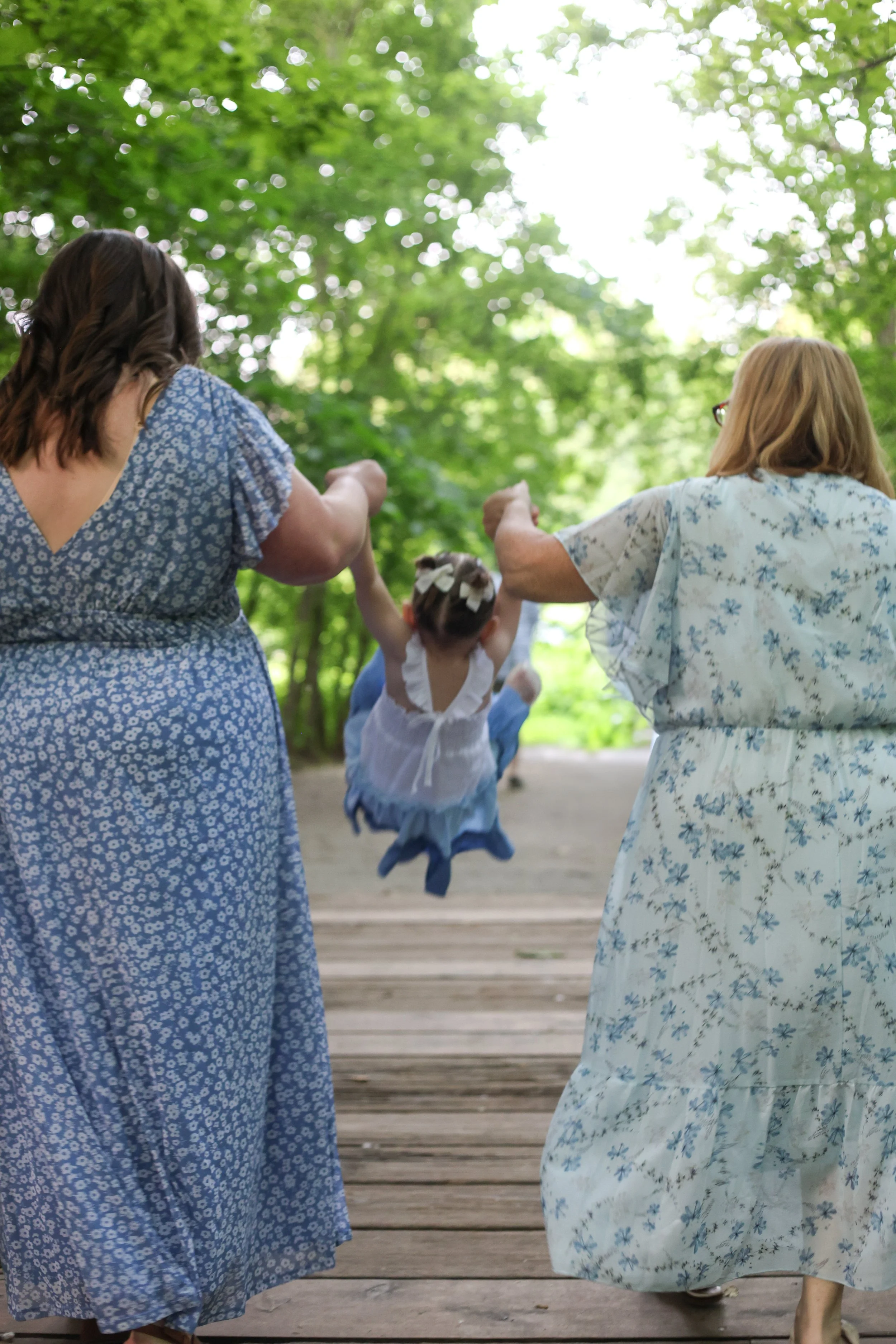 Two women and a young girl are outside on a wooden path passing a lush green area with trees. The women are holding the girl by her hands as she is swinging between them. All are dressed in light blue and white floral dresses.