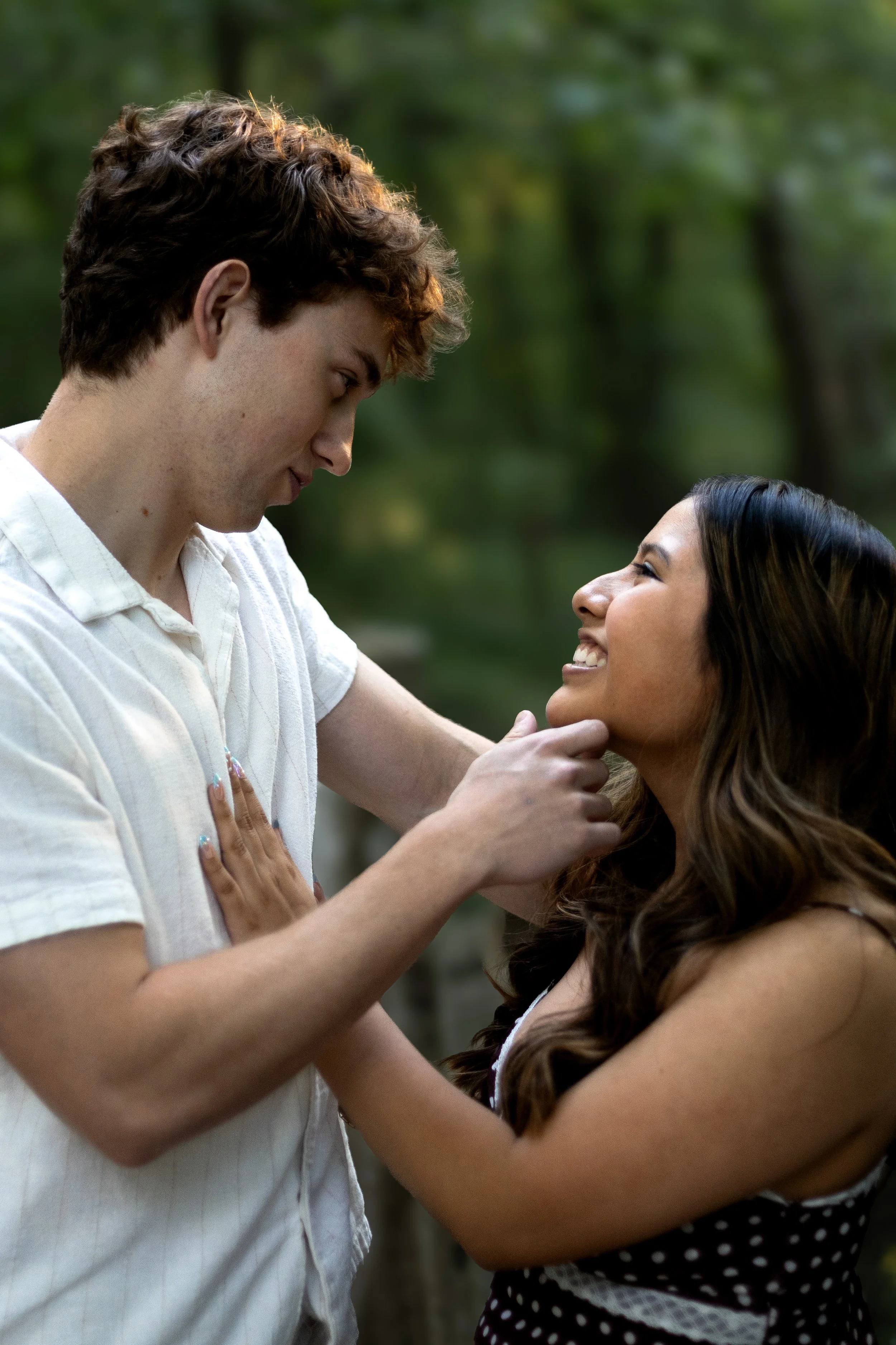 A young couple sharing a moment outdoors. The man, with curly hair and wearing a white shirt, is gazing into the woman's eyes. The woman has long dark hair, wearing a polka-dot dress, smiling with her hand on her chest. They are surrounded by blurred