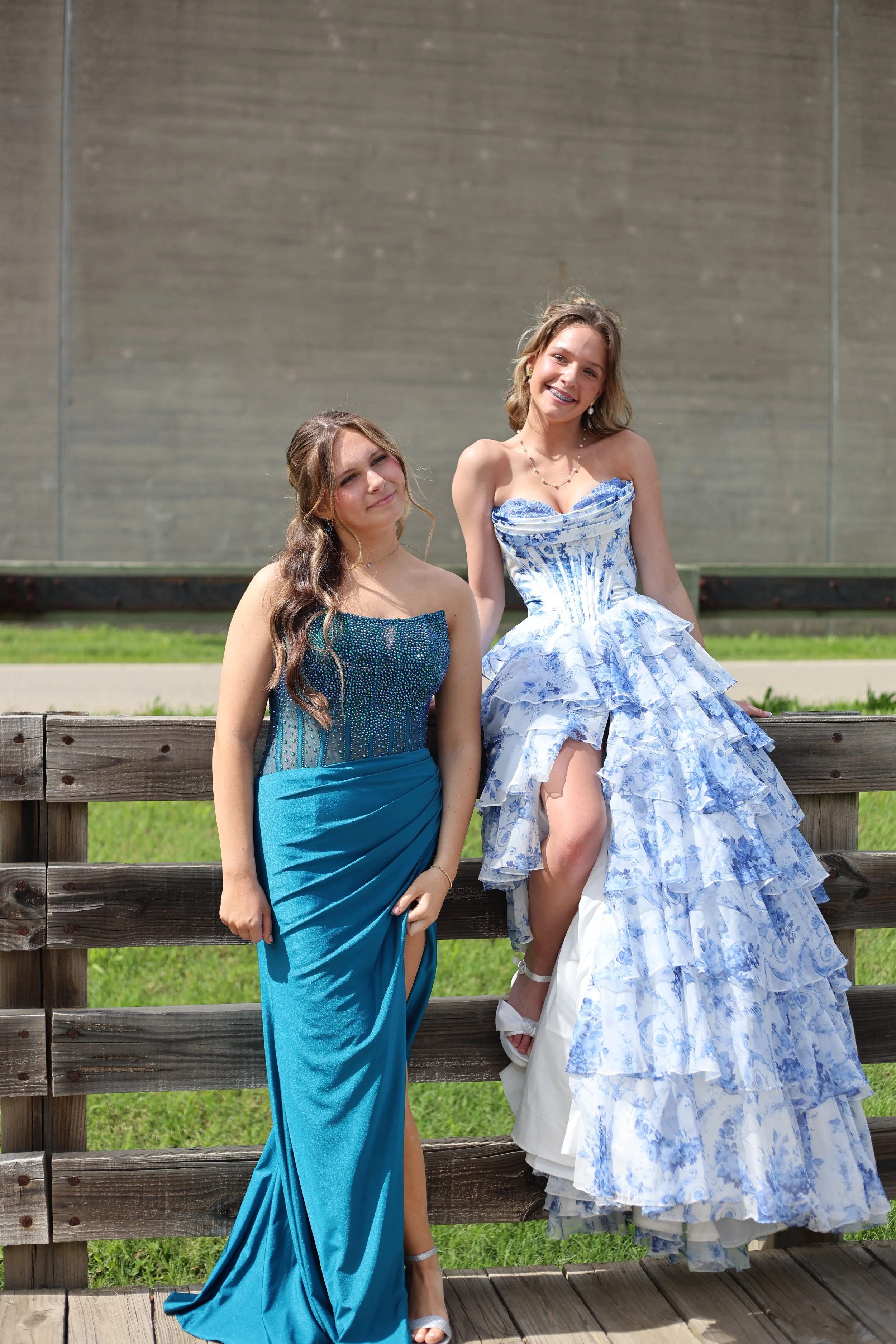 Two young women posing on a wooden fence outdoors, dressed in formal gowns, with a plain wall in the background.