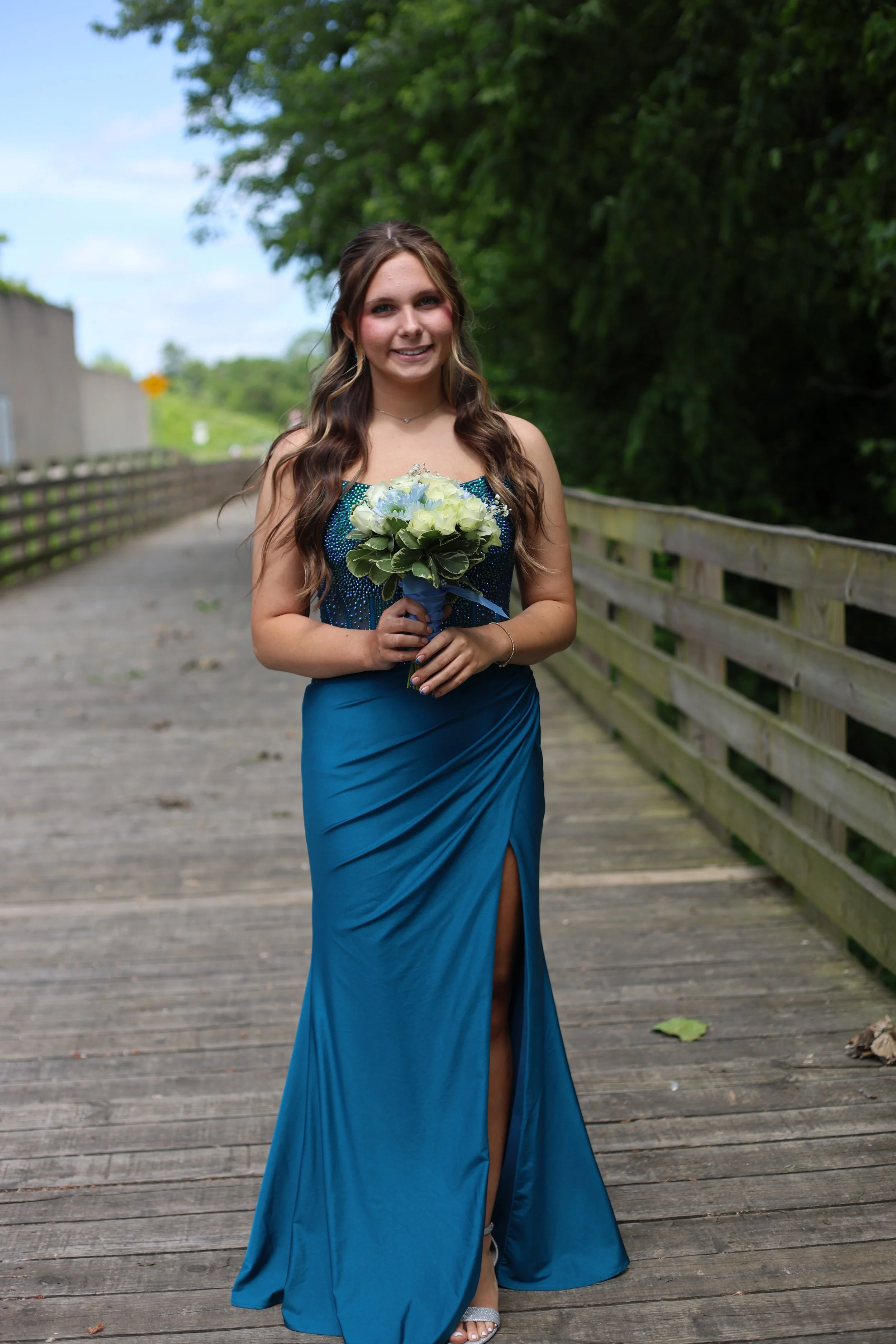A young woman wearing a long, elegant, blue evening gown with a high slit and fitted bodice, standing on a wooden bridge, holding a bouquet of white and blue flowers, smiling at the camera, with lush green trees in the background.