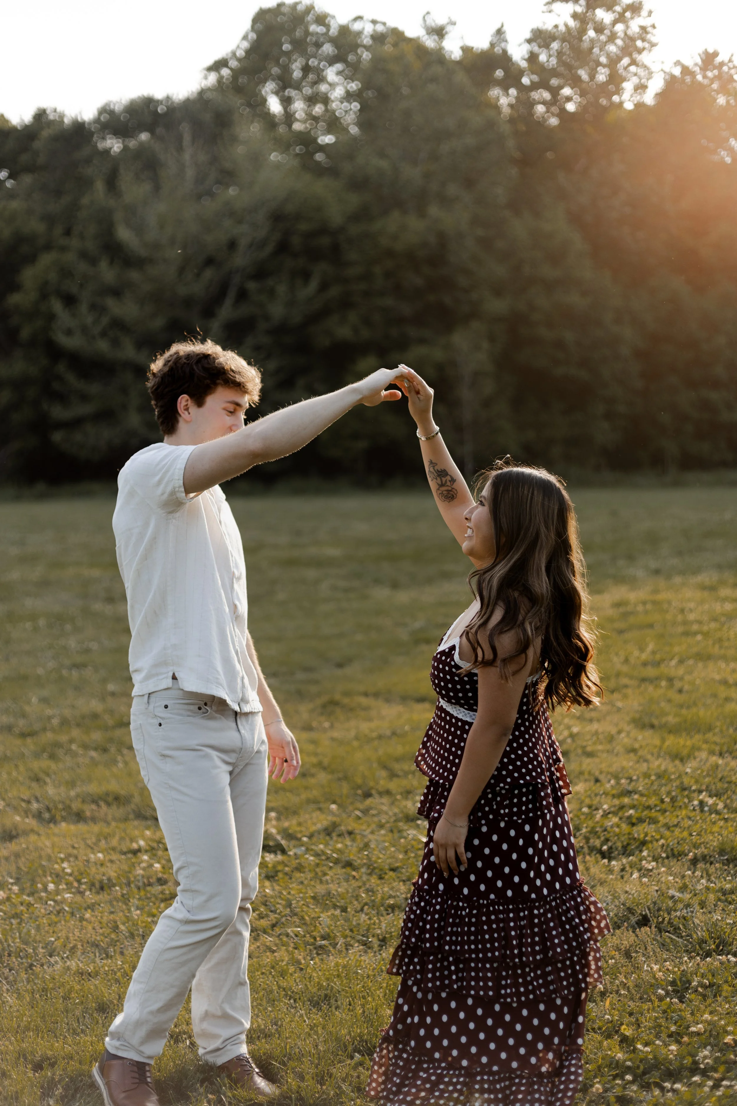 A young man and woman standing outdoors, holding hands and smiling at each other during sunset in a grassy field with trees in the background.