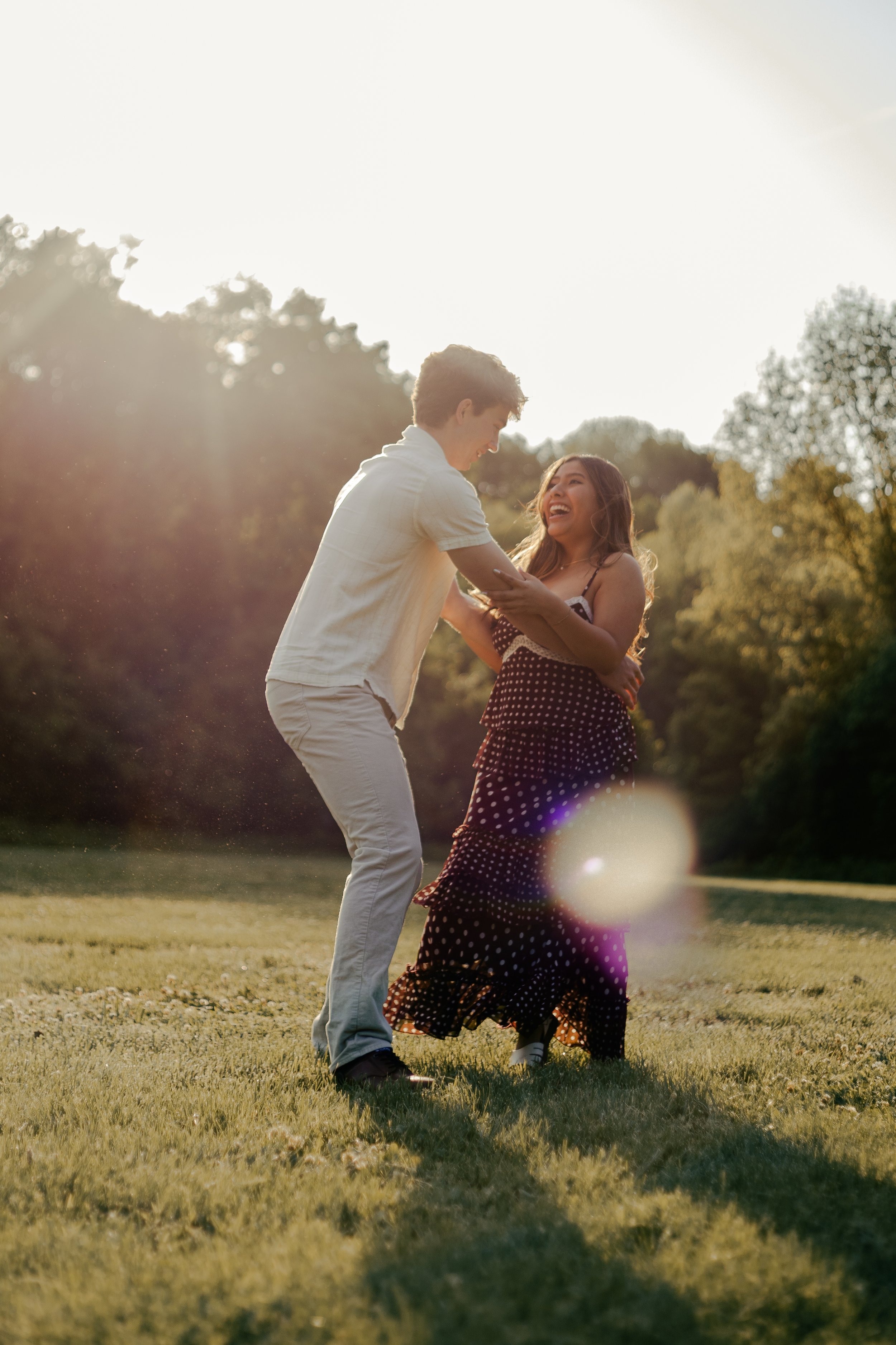 A young couple is outdoor on a grassy field during sunset, smiling and looking at each other, with trees in the background.