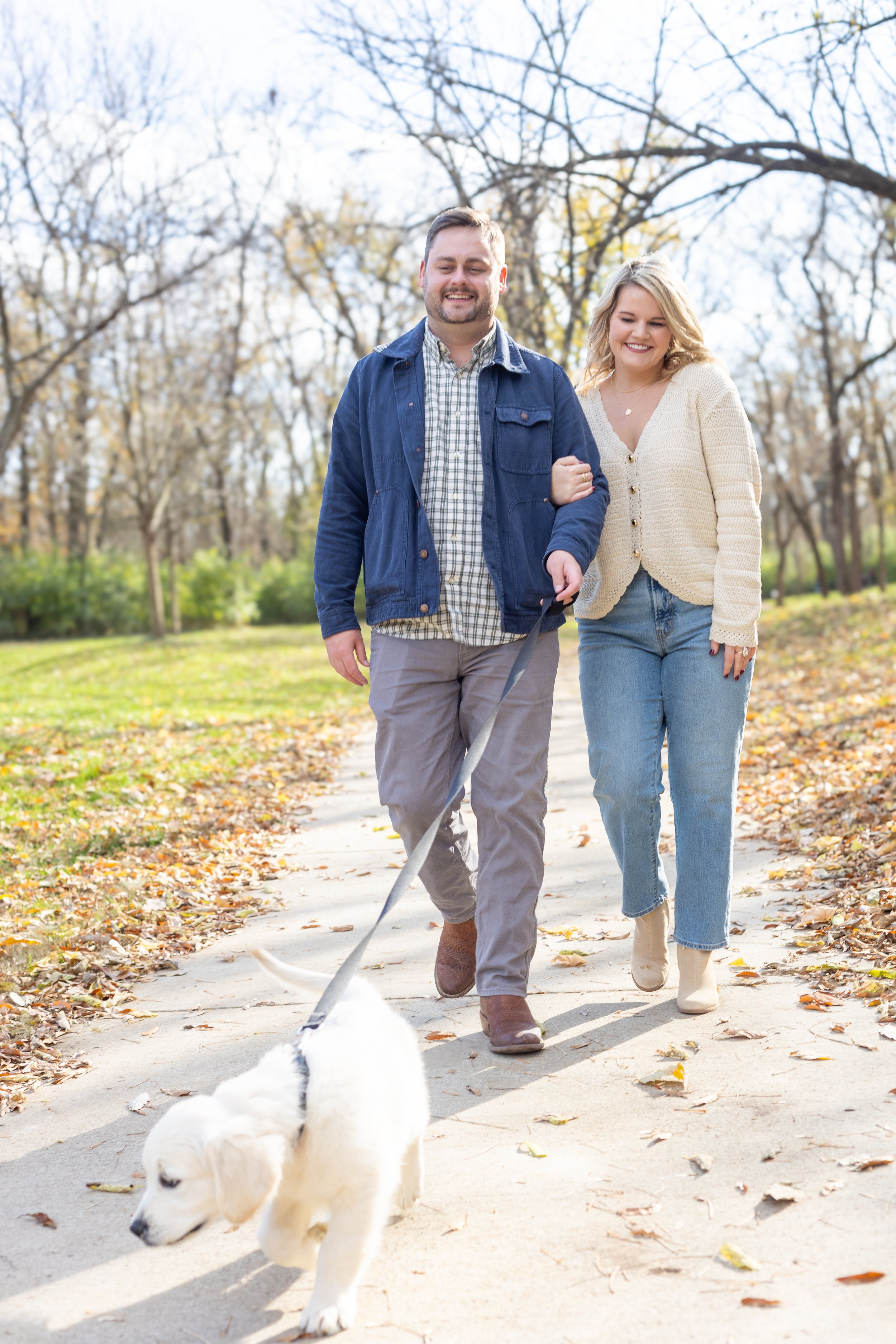 A couple walks arm-in-arm outdoors on a fall day, smiling. They are walking a large white dog with black markings on a leash along a leaf-covered path with trees in the background.