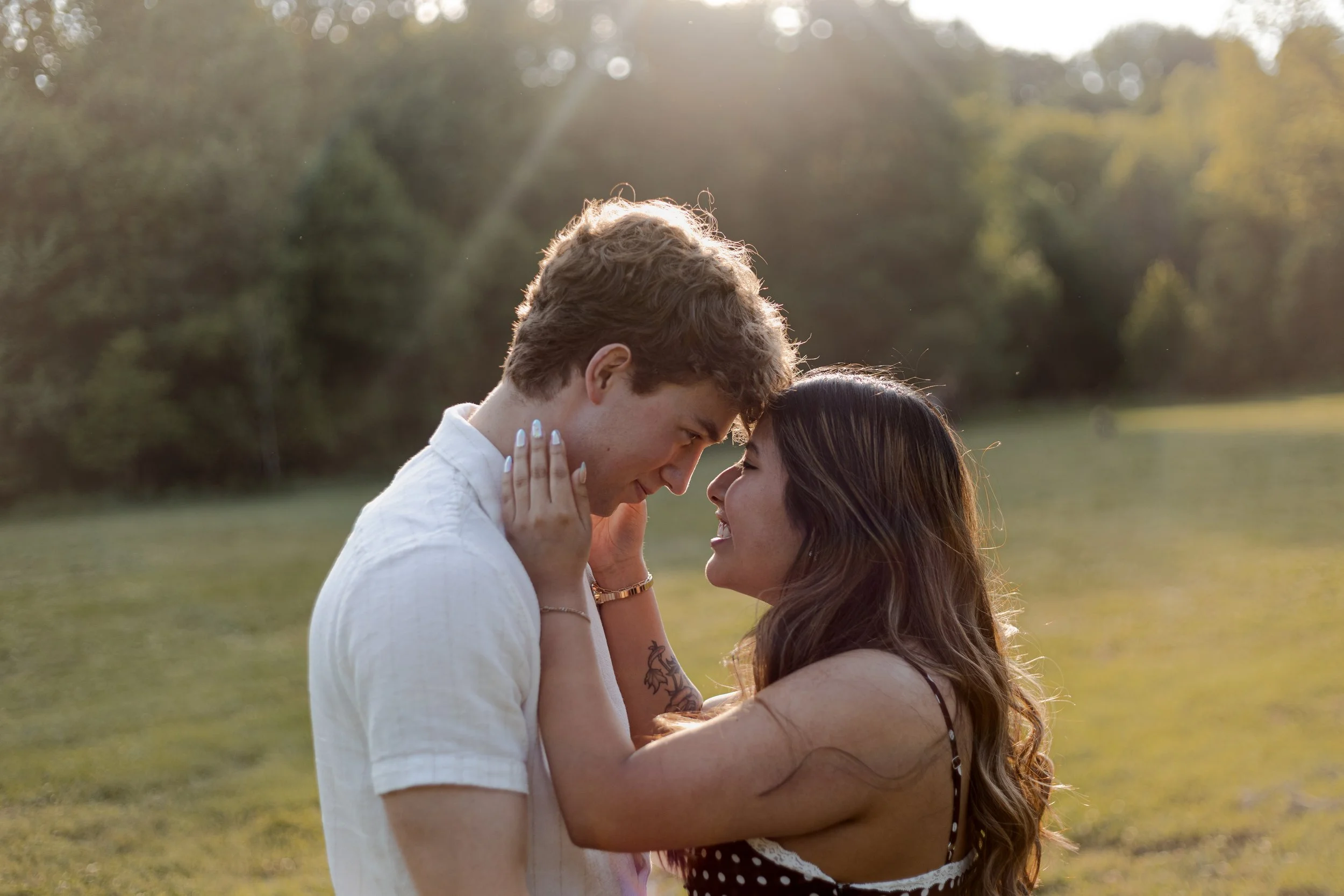 A young man and woman standing close together outdoors during sunset, touching foreheads and smiling with eyes closed, in a park with trees in the background.