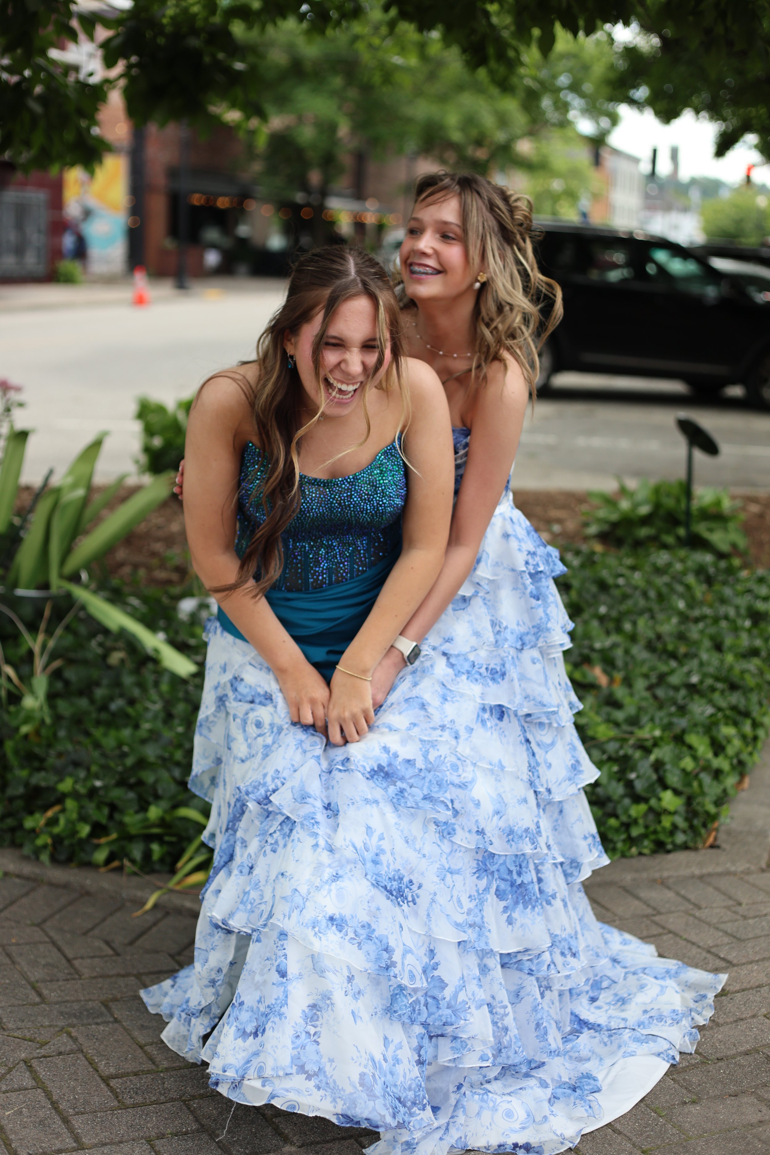 Two young women in blue and white dresses sharing a joyful moment outdoors on a city street, surrounded by greenery and parked cars.