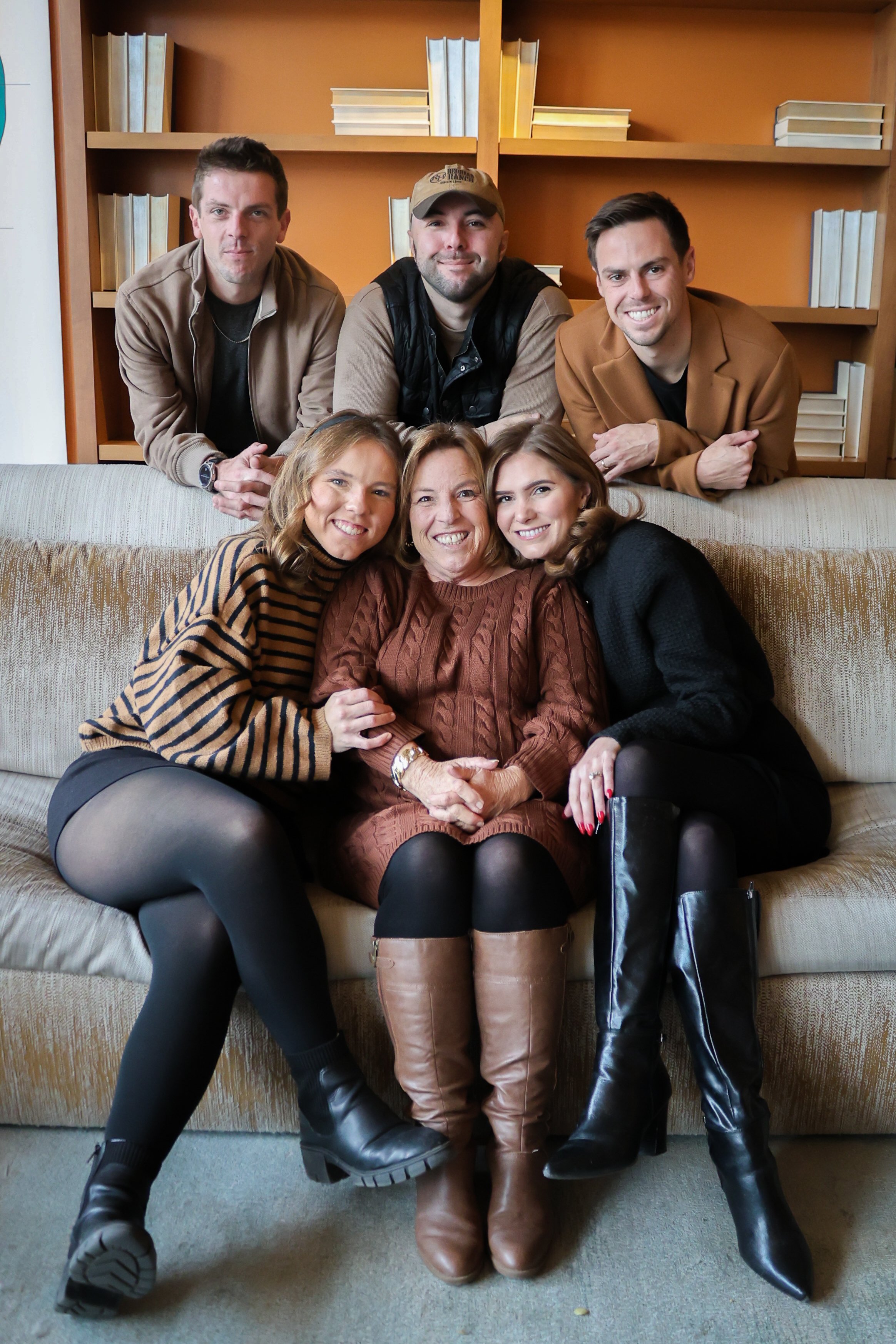 Group of five people, three men and two women, sitting on a beige sofa in front of a wooden bookshelf. The women are sitting on the floor, and the men are leaning on the back of the sofa. All are smiling and looking at the camera.