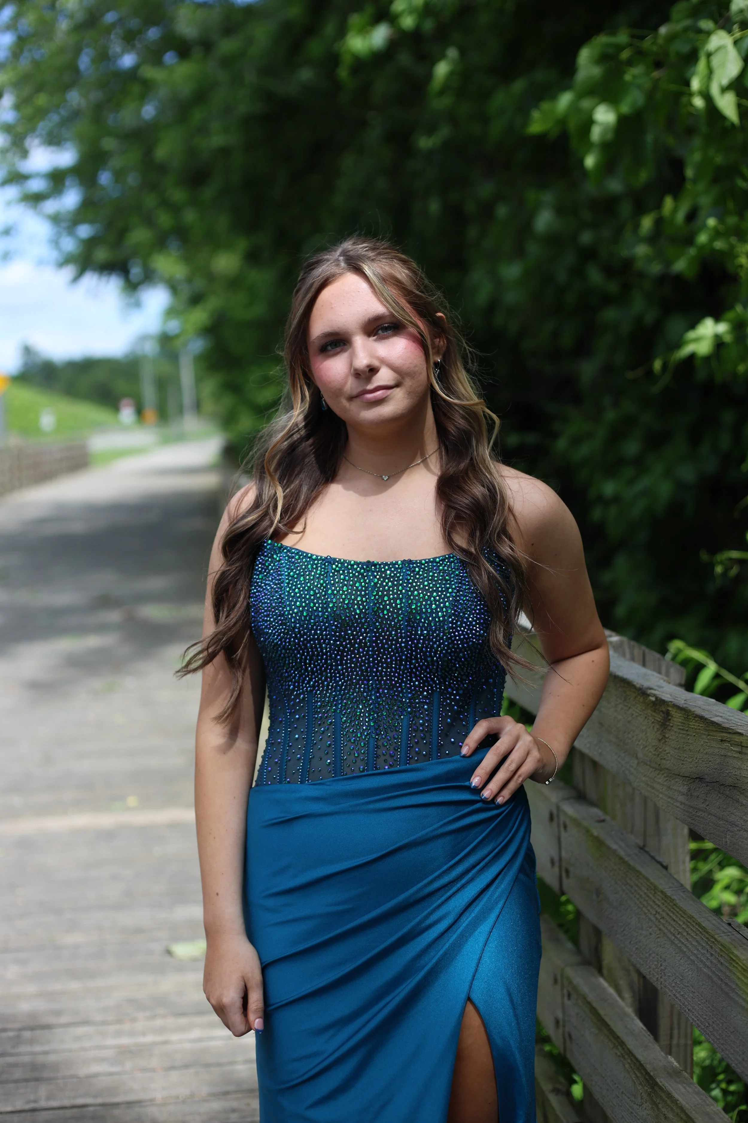 Young woman in a blue evening gown standing outdoors on a wooden pathway, with green trees and a wooden railing behind her.
