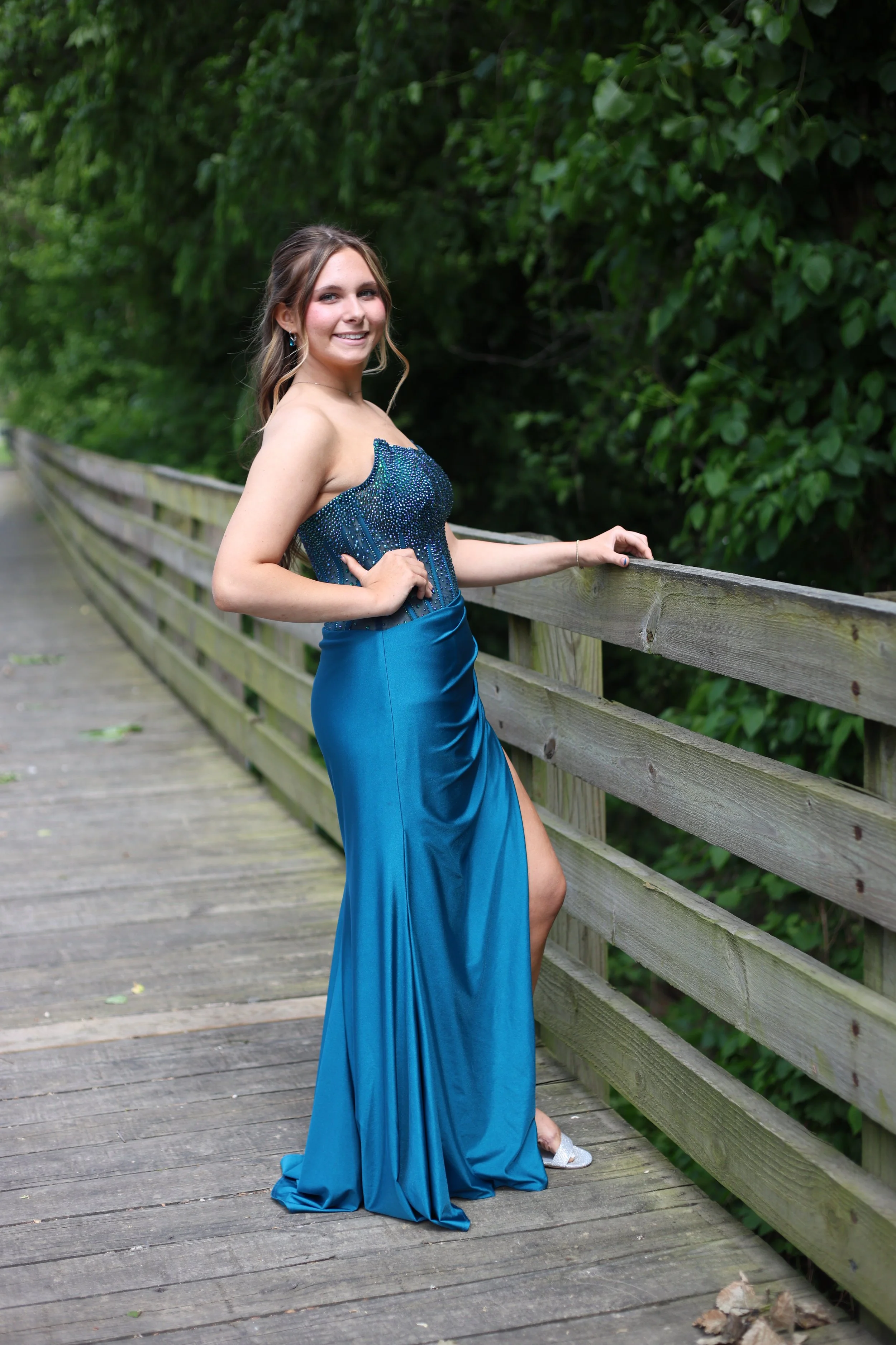 Young woman in a strapless, teal evening gown standing on a wooden bridge with greenery in the background.