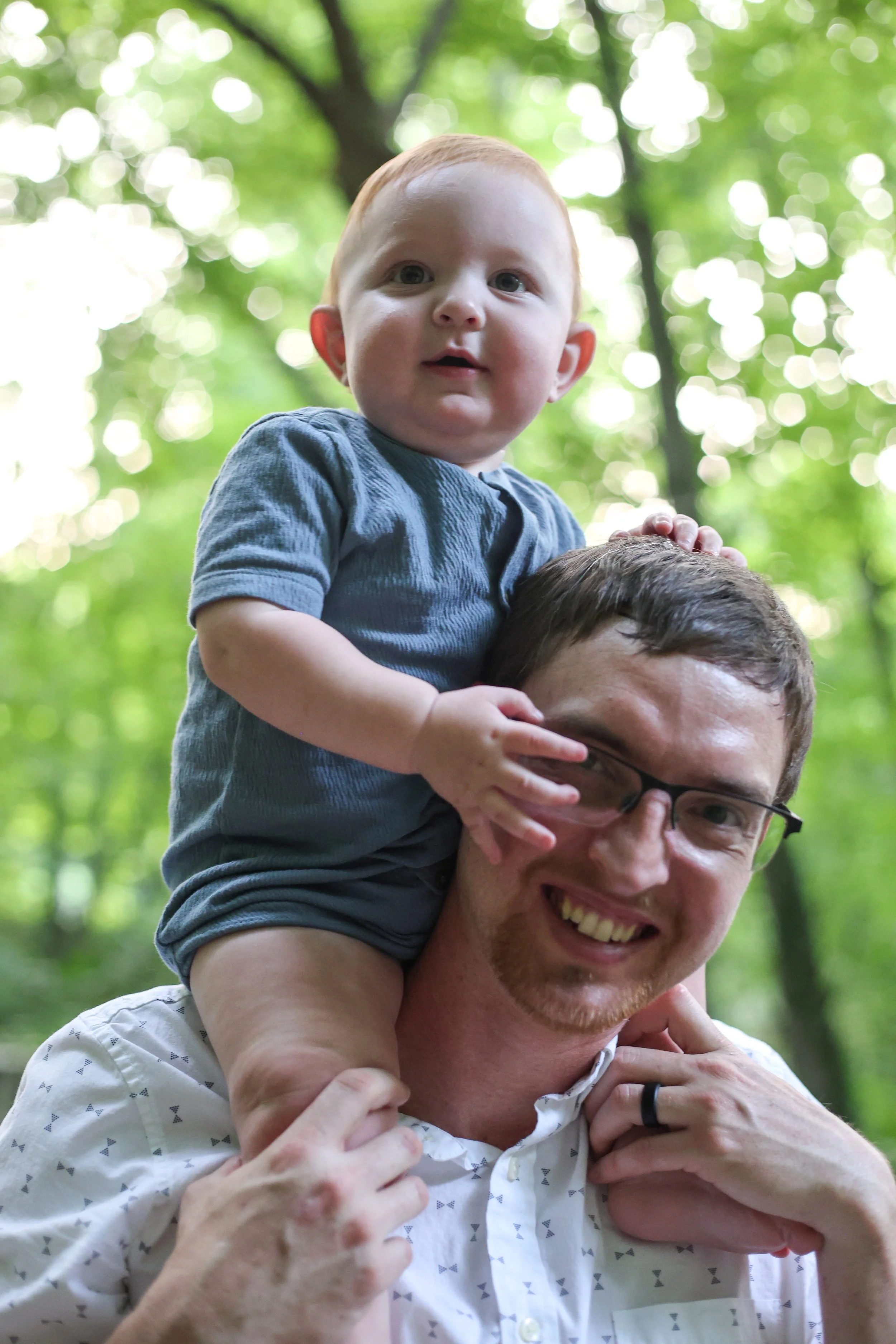 A man with glasses smiling and carrying a young child on his shoulders in a wooded area with green foliage and sunlight filtering through the trees.