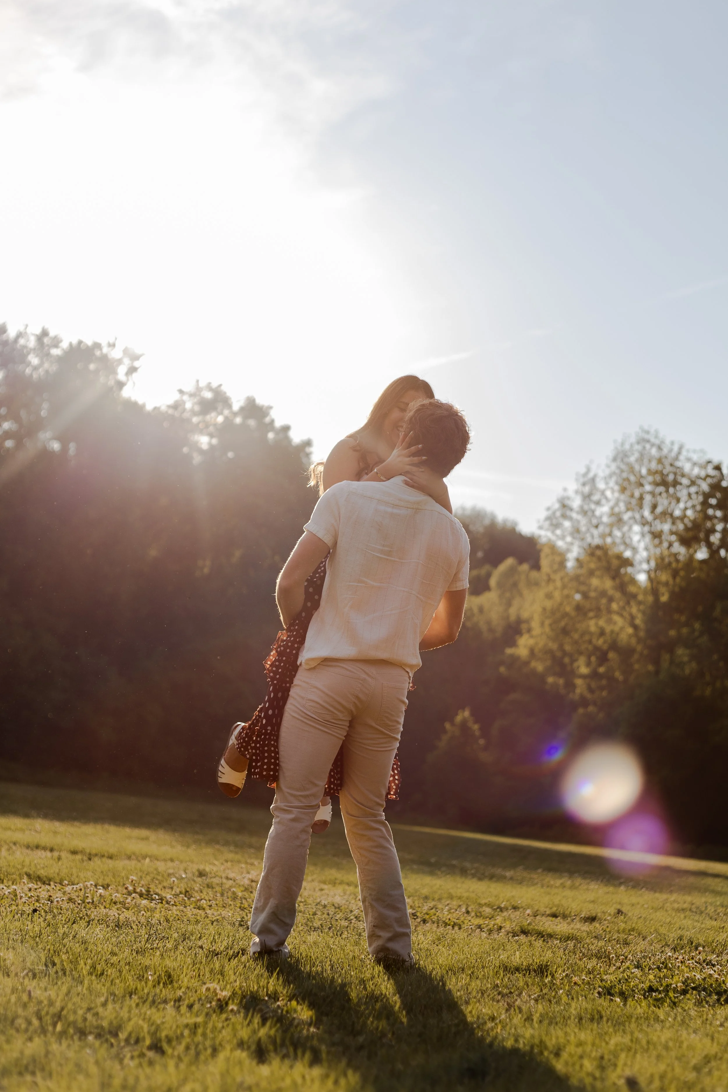 A couple in a park during sunset, with the woman sitting on the man's shoulders, smiling and touching his face, with trees and a grassy field in the background.