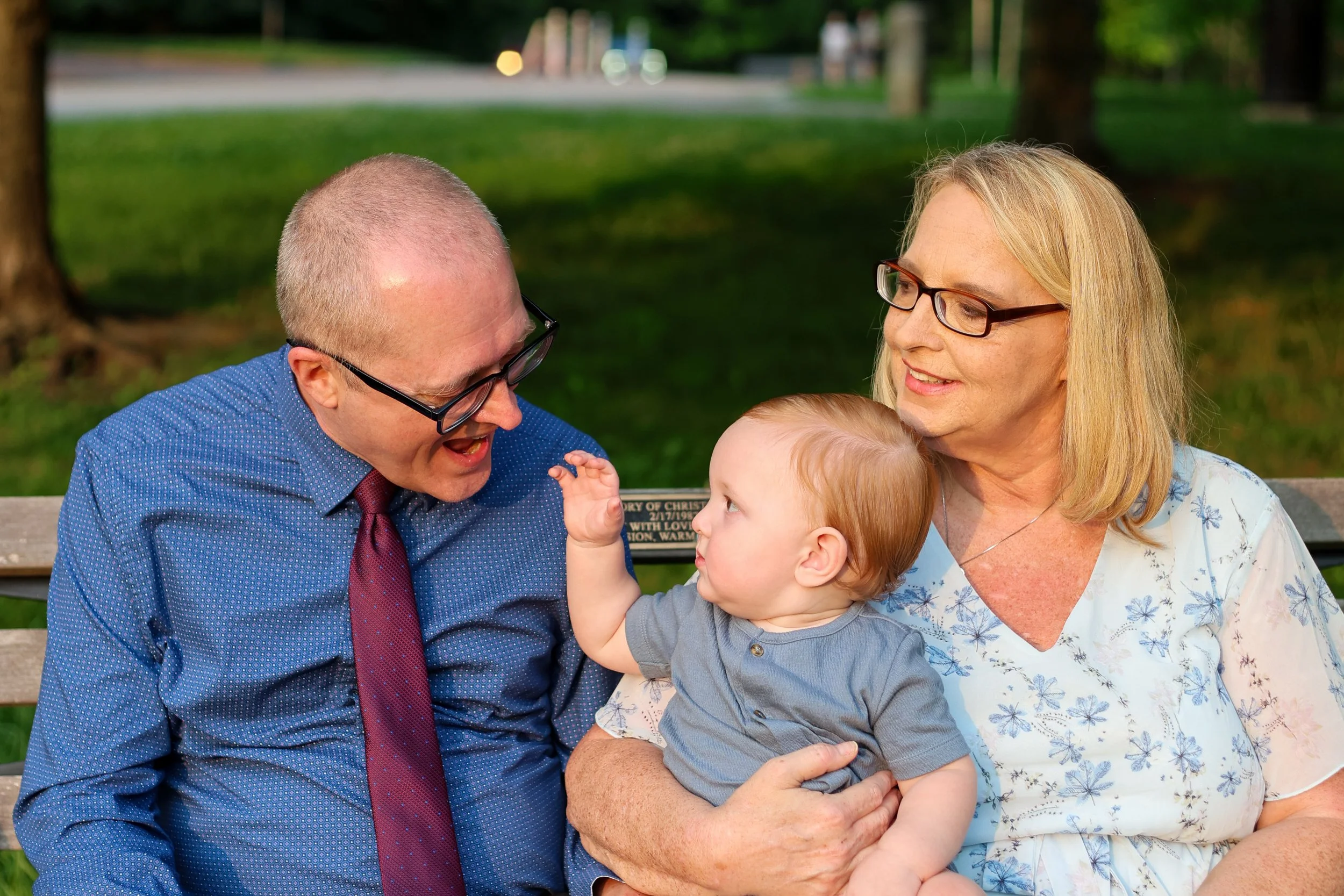 A man, woman, and a young child sit on a park bench outdoors surrounded by trees. They are smiling and engaging with each other, with the man and woman looking at the child who is reaching towards the man.