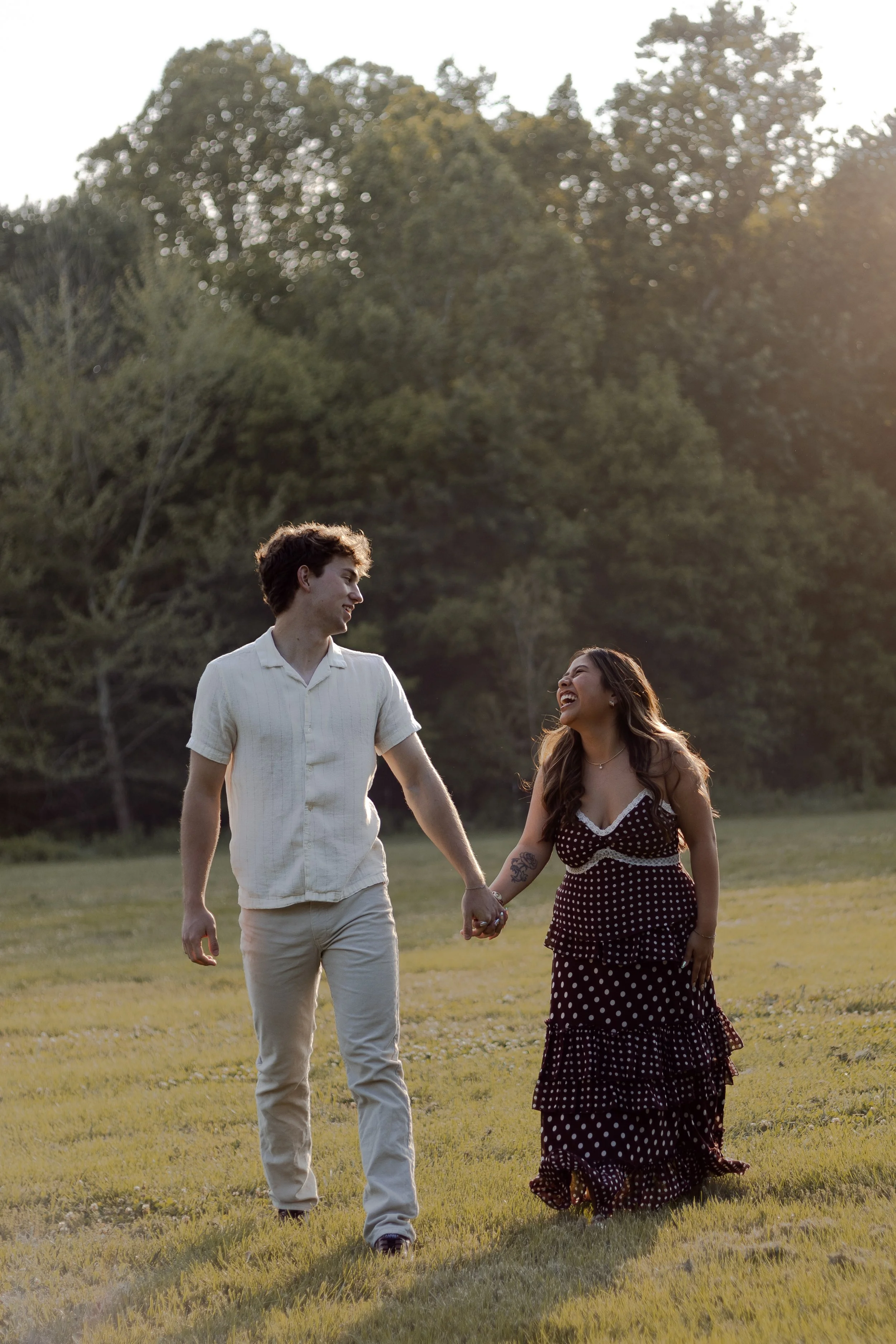 A young couple holding hands and walking through a grassy field with trees in the background, during sunset.