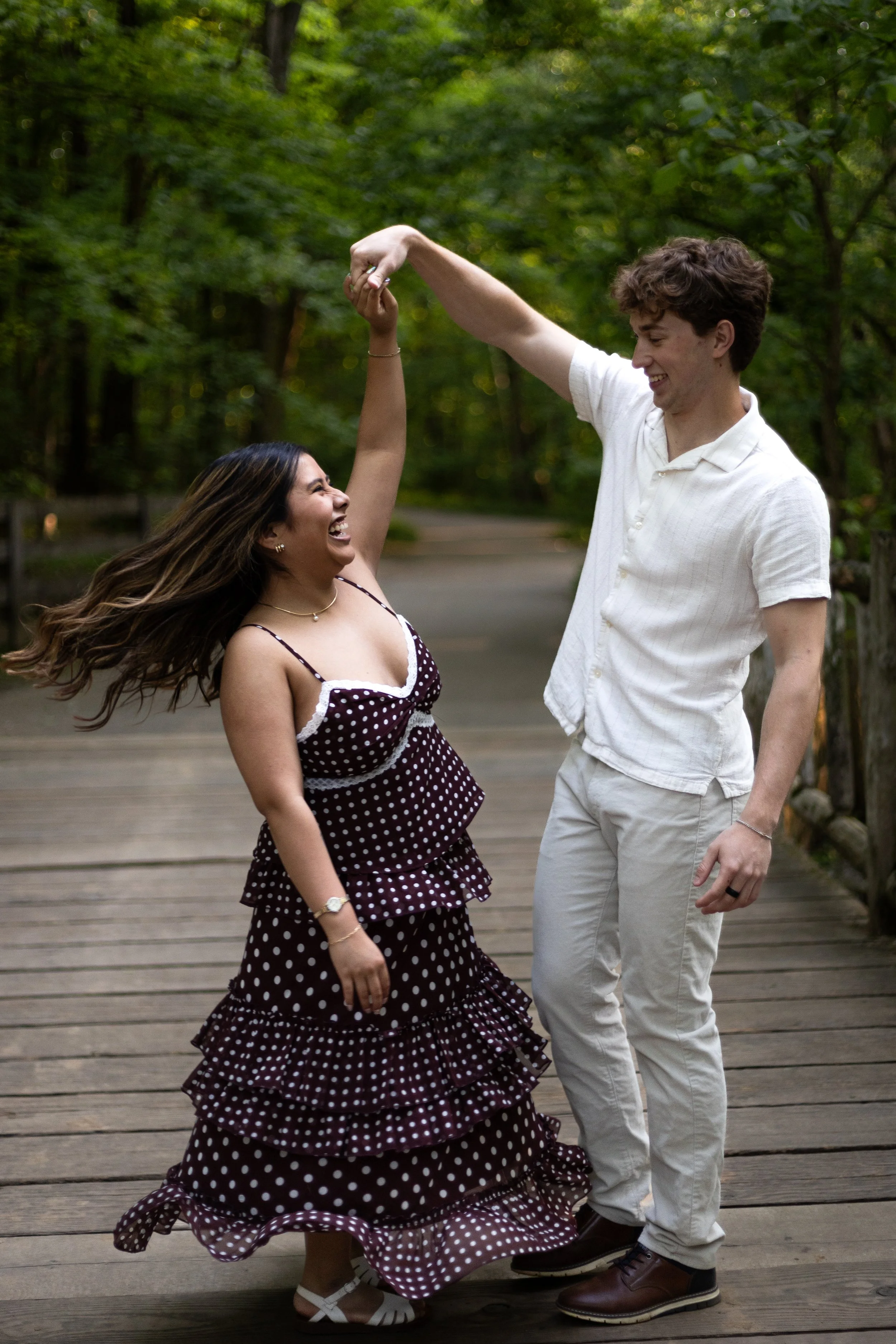 A young woman in a polka dot dress and a young man in a white shirt dance together on a wooden bridge surrounded by green trees, smiling and enjoying the moment.