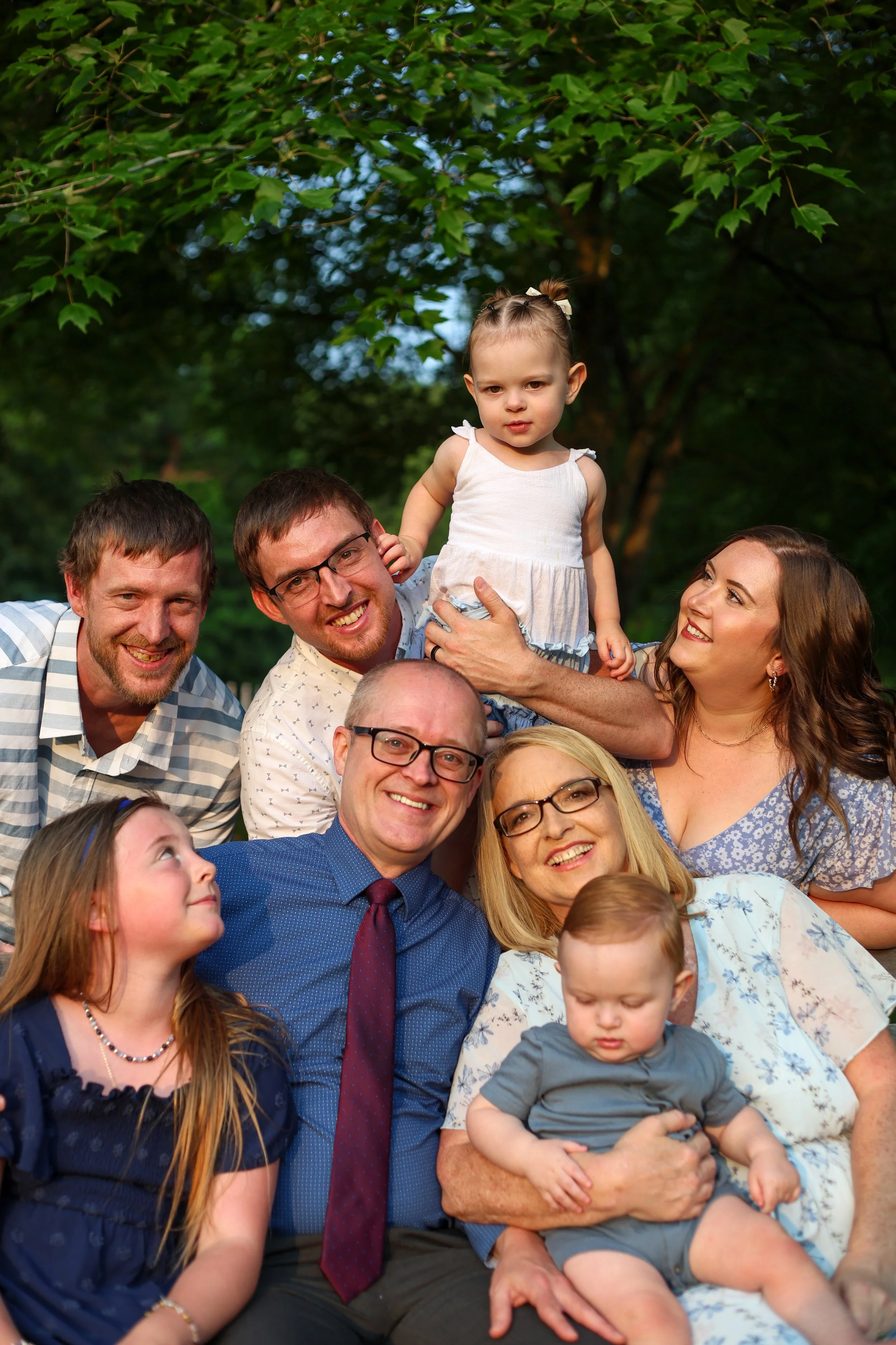 A group of people, including several adults and children, gathered outdoors in a park or garden with trees and greenery in the background. They are smiling and posing for a photo, with some children being held by adults.