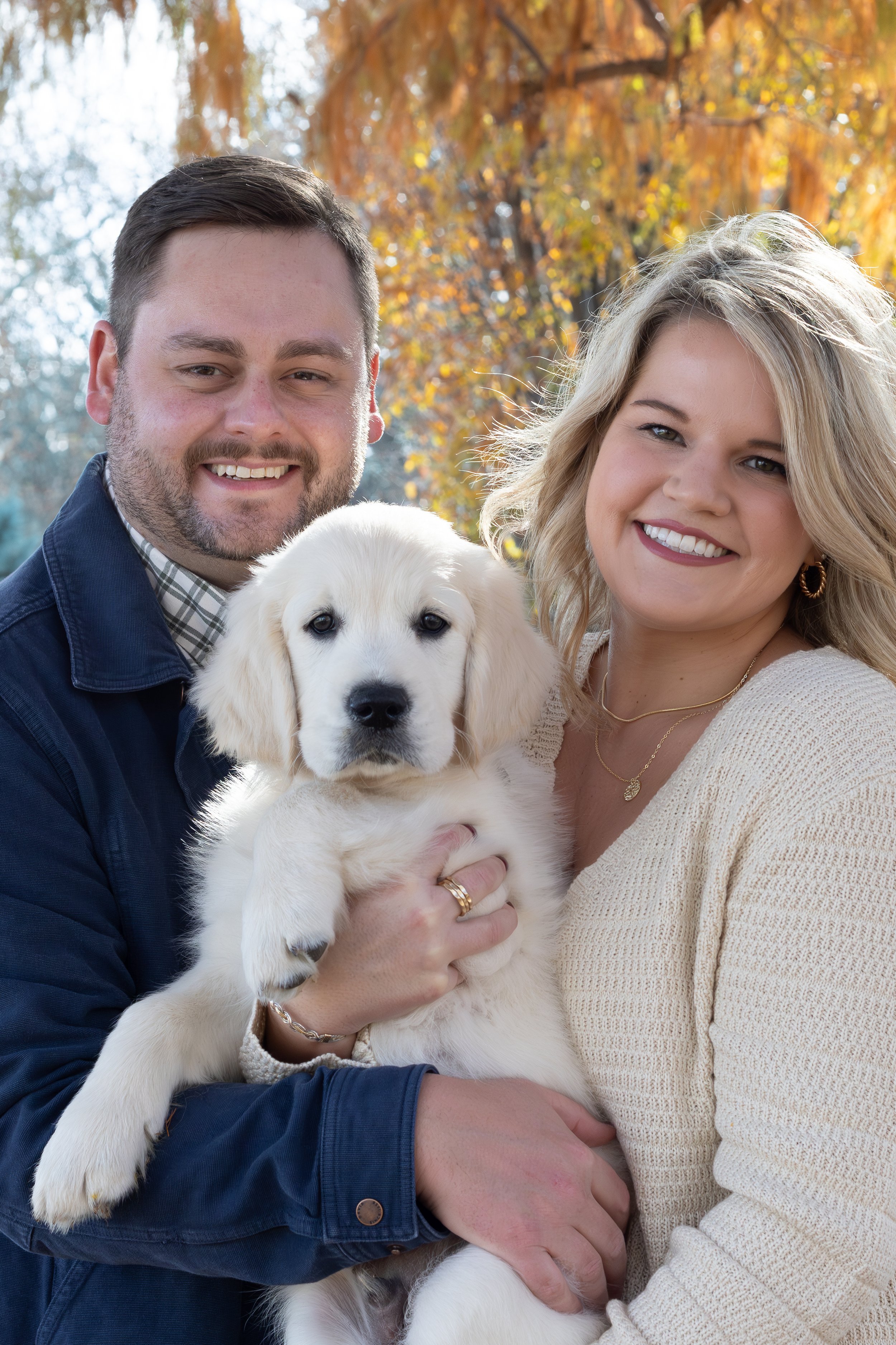 A smiling man and woman holding a golden retriever puppy outdoors with autumn trees in the background.