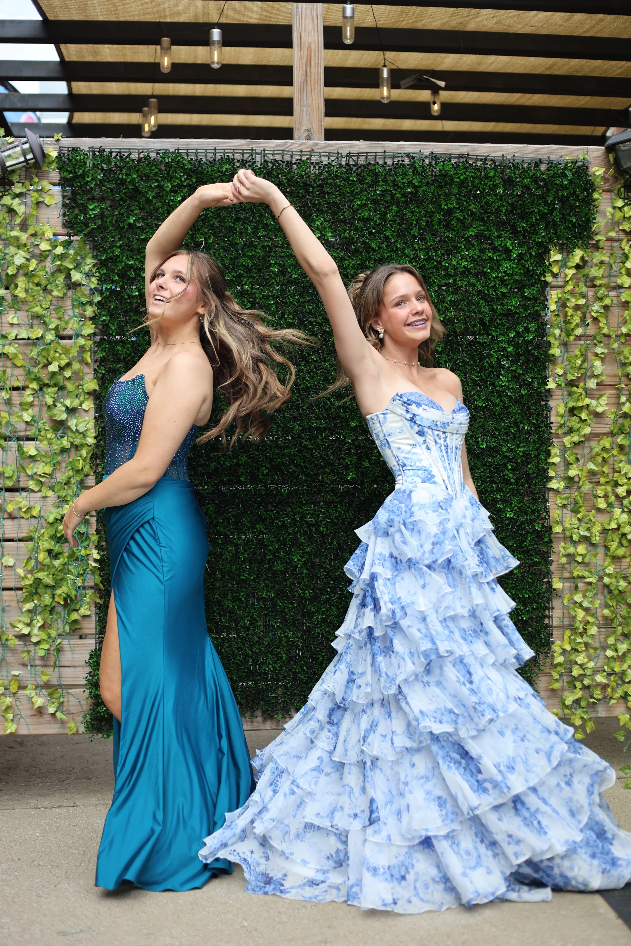 Two young women in elegant blue dresses dancing together outdoors in front of a green hedge wall.