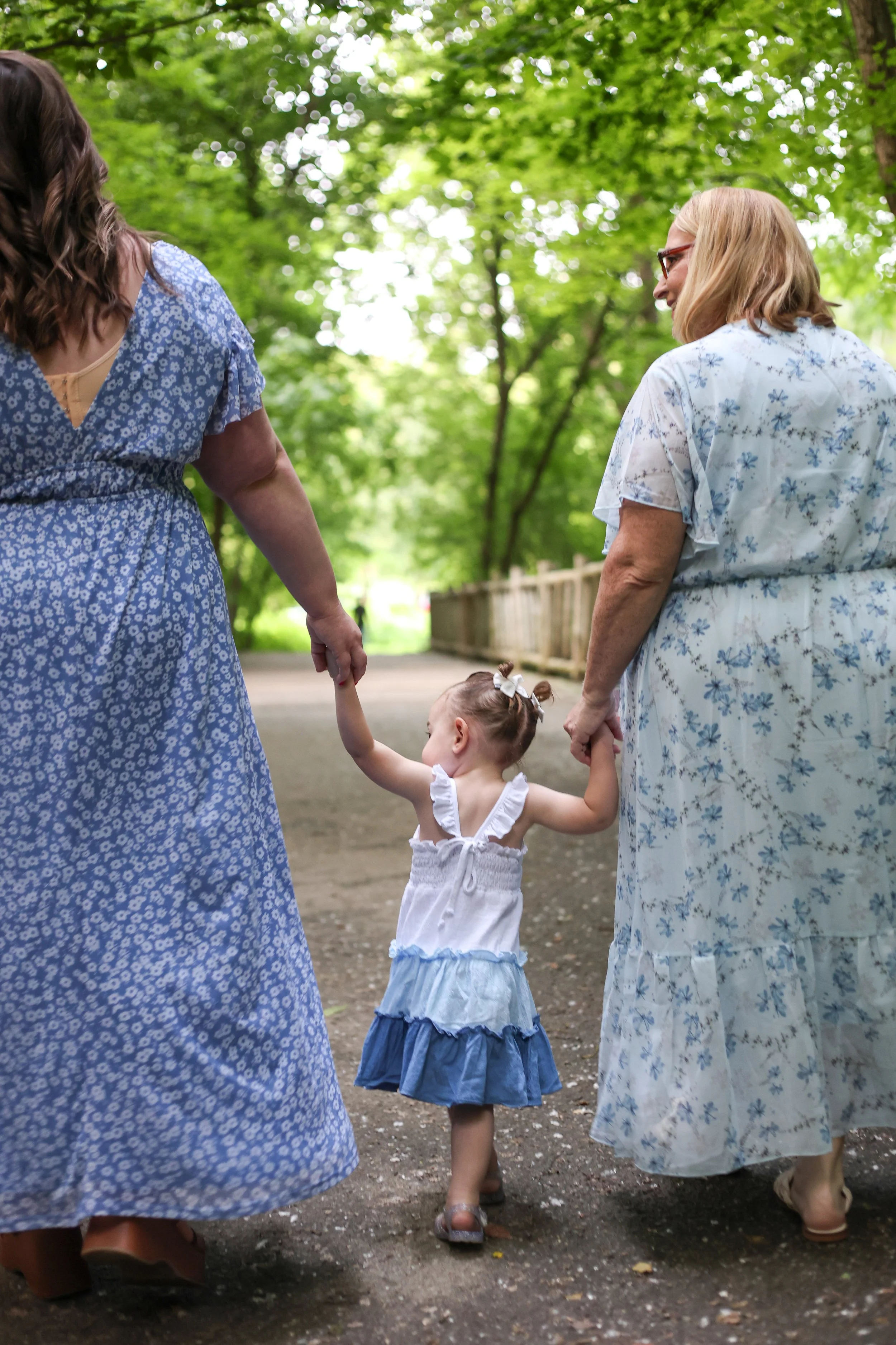 Two women holding the hands of a young girl in a blue and white dress, walking on a dirt path in a green park with trees.