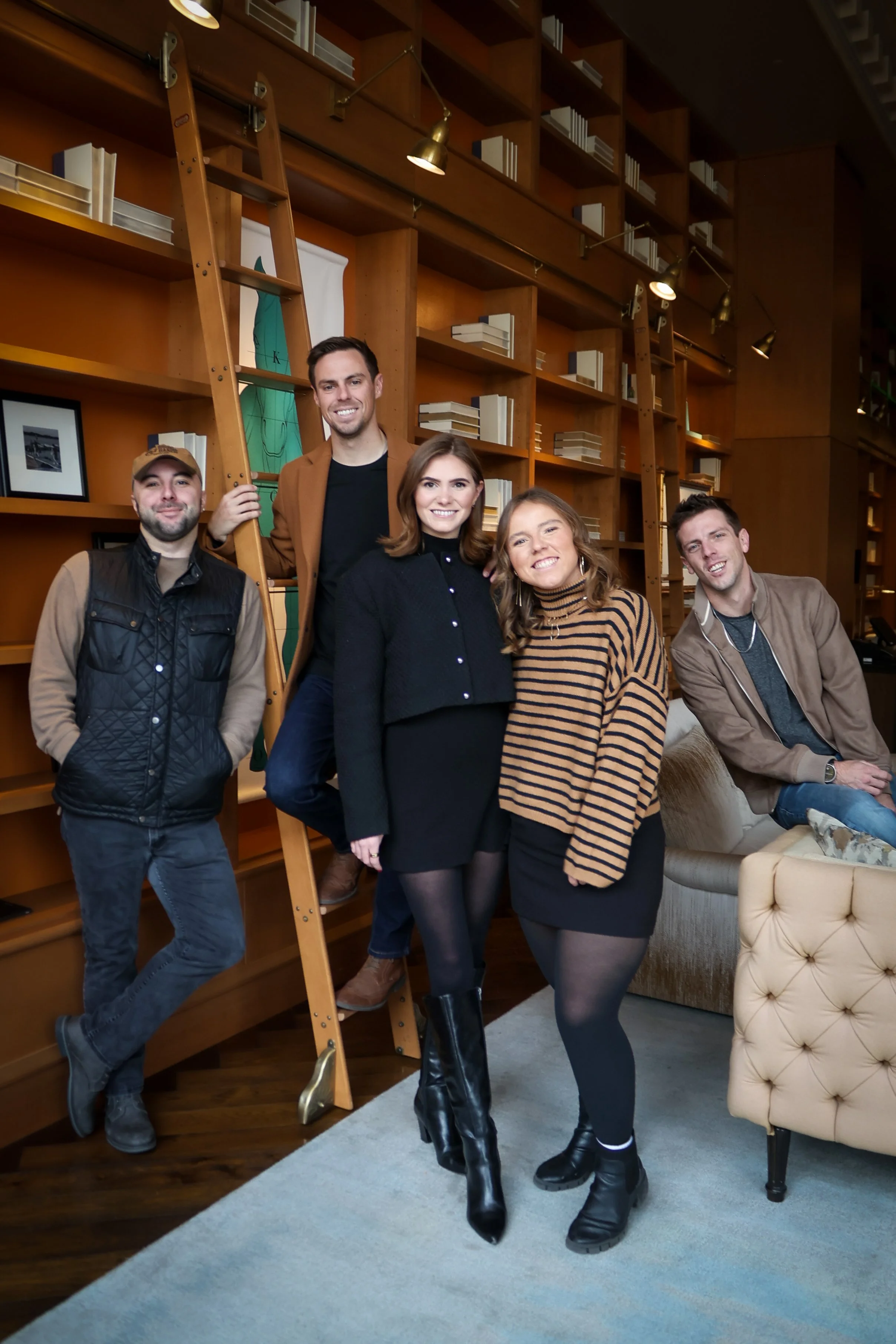Group of five people posing indoors in front of a wooden bookshelf, with two ladders leaning against it, in a cozy room with framed pictures and a beige tufted chair.