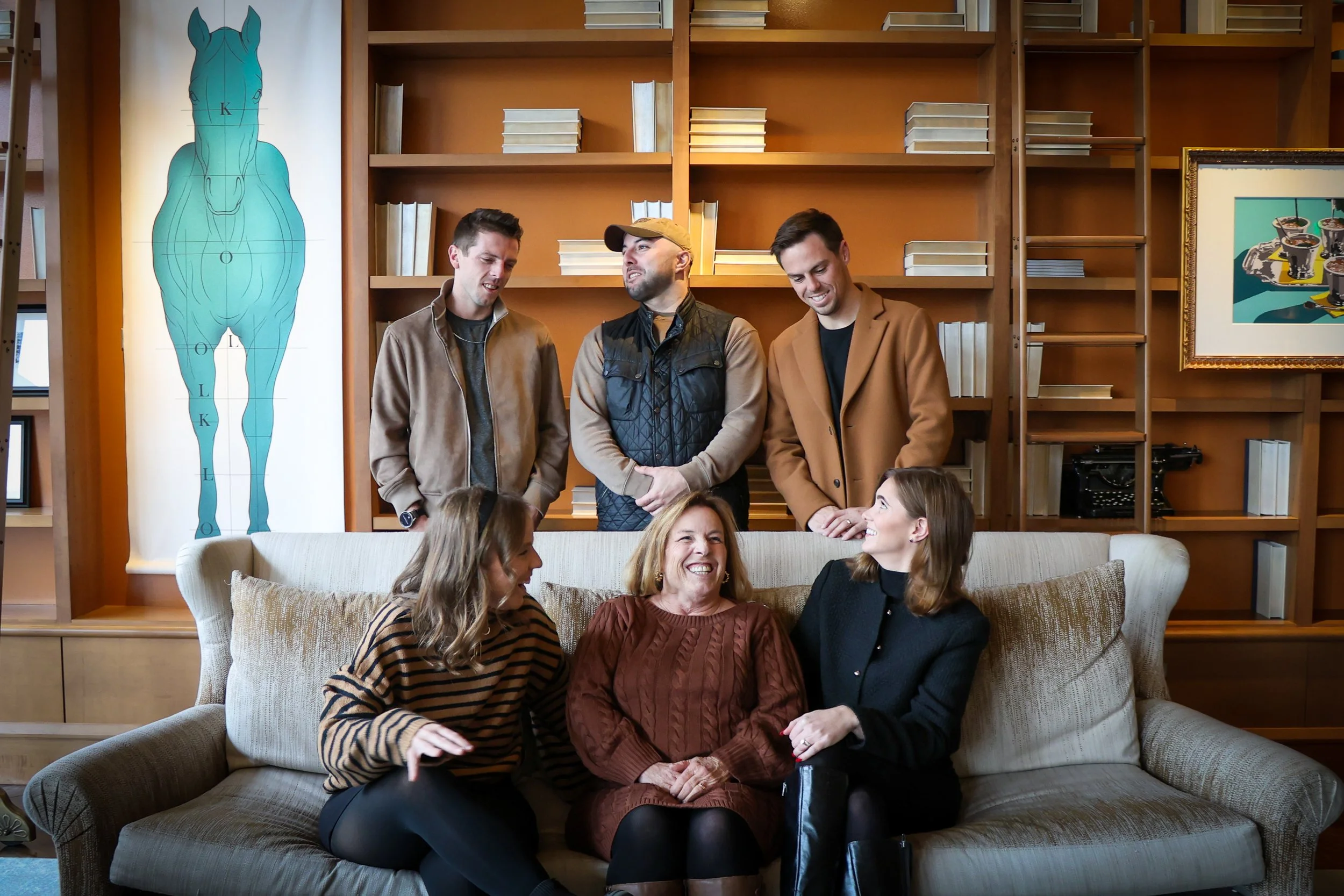 A group of people sitting on a couch and smiling, with three men standing behind them, in a cozy room with wooden shelves and artwork including a large anatomical poster of a horse.